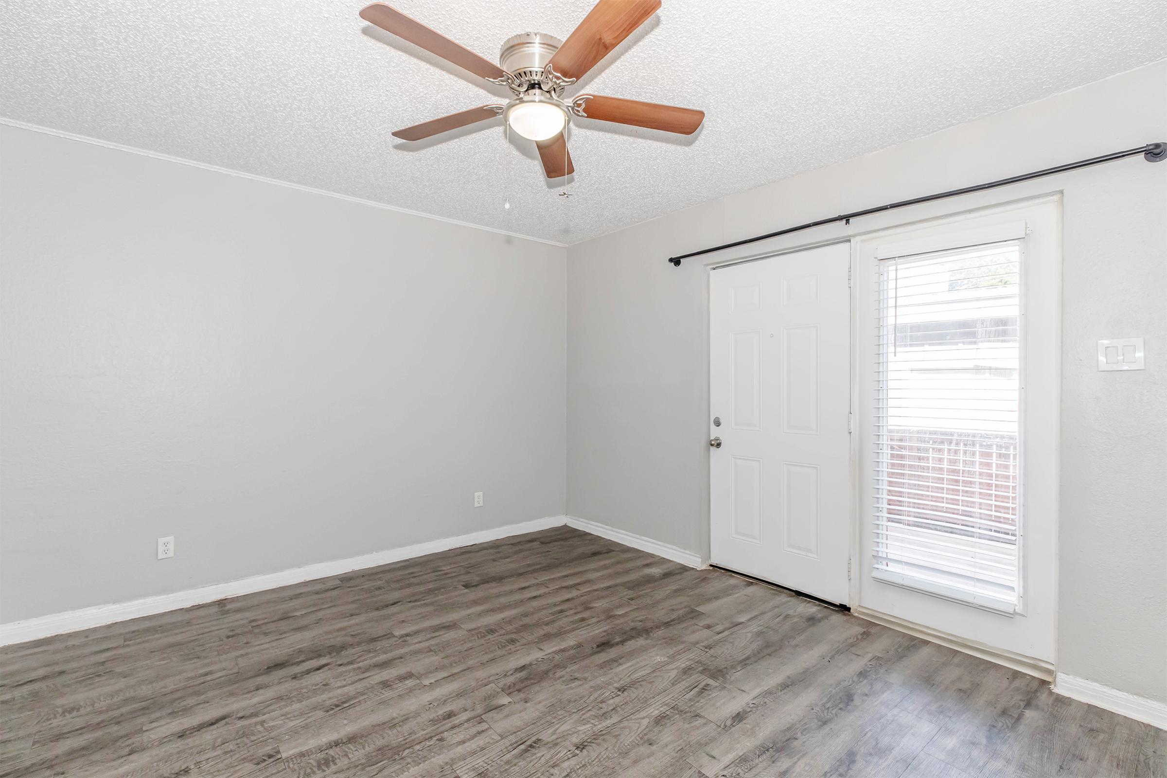 A small, empty room with light gray walls and a ceiling fan. There is a closed door on the left and a window on the right, allowing natural light to enter. The floor is covered in laminate wood, providing a clean and modern appearance.