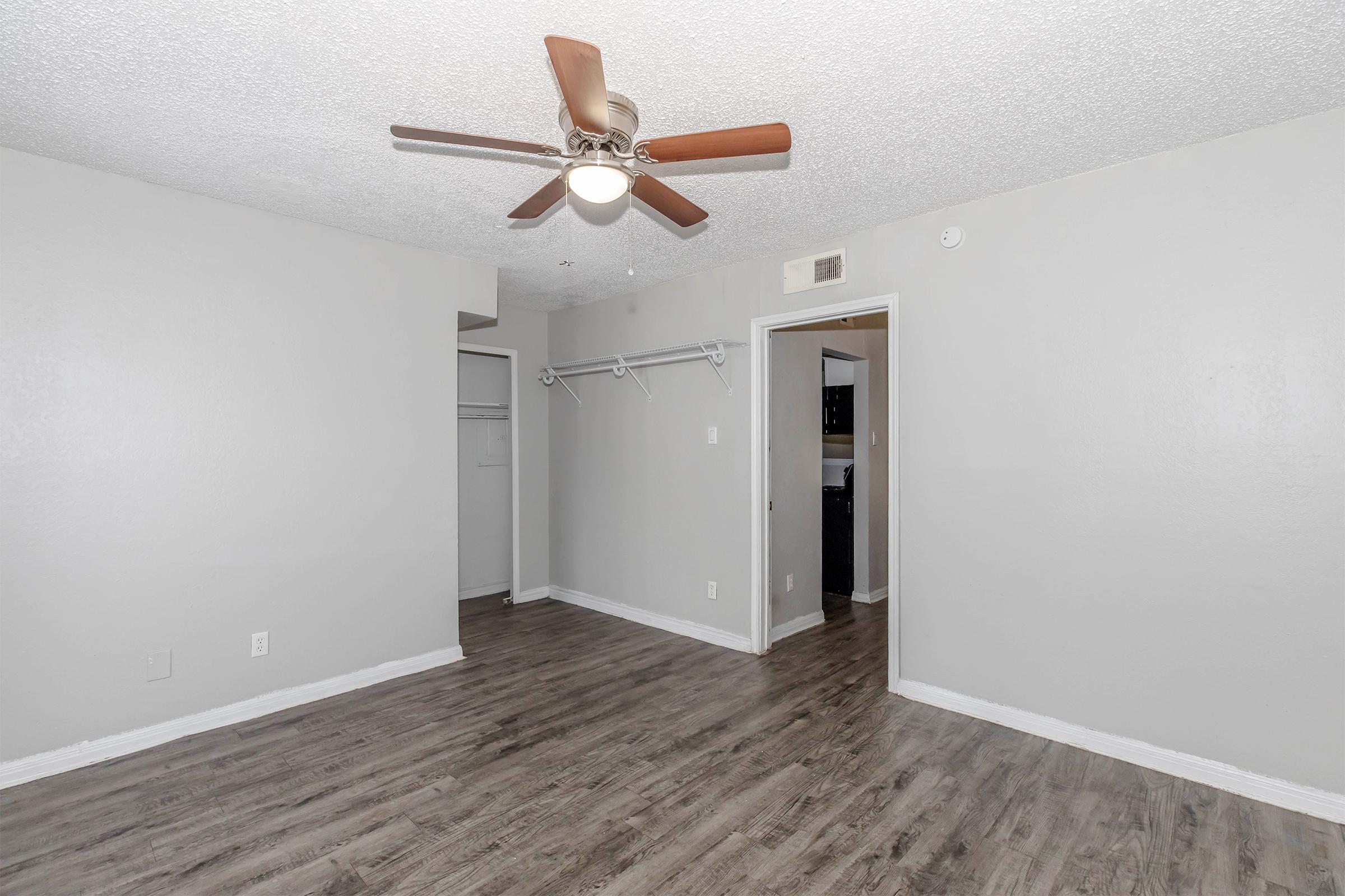 A spacious, empty room with light gray walls and a ceiling fan. The flooring is a laminate wood style. There is a closet space on the left with a hanging rod, and a doorway leading to another room on the right. Overall, the room is well-lit and ready for furnishing.