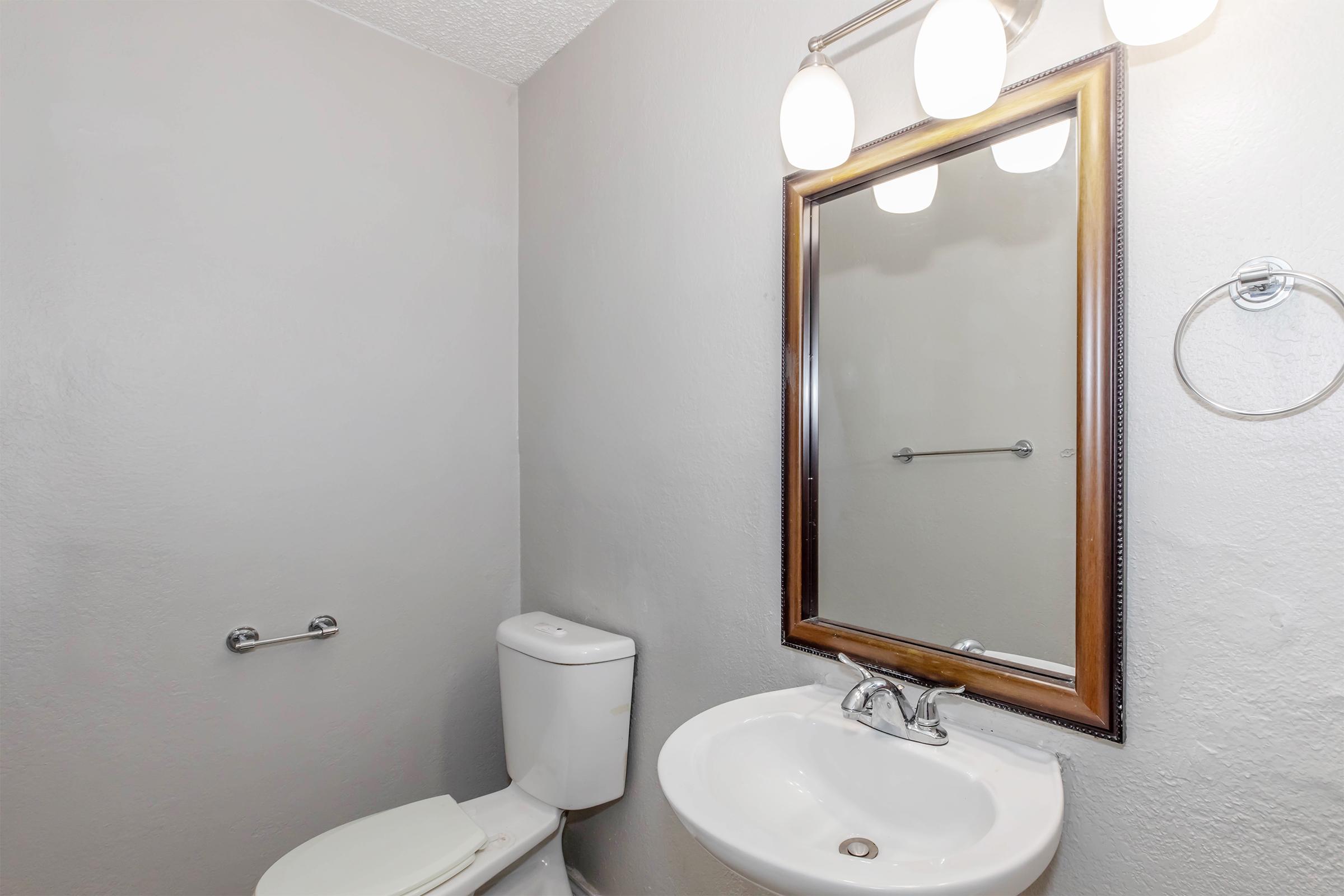 A clean, well-lit bathroom featuring a white toilet, a small sink with a faucet, and a round mirror above the sink. There are two light fixtures mounted above the mirror, and a towel ring is attached to the wall. The walls are painted a light gray color.