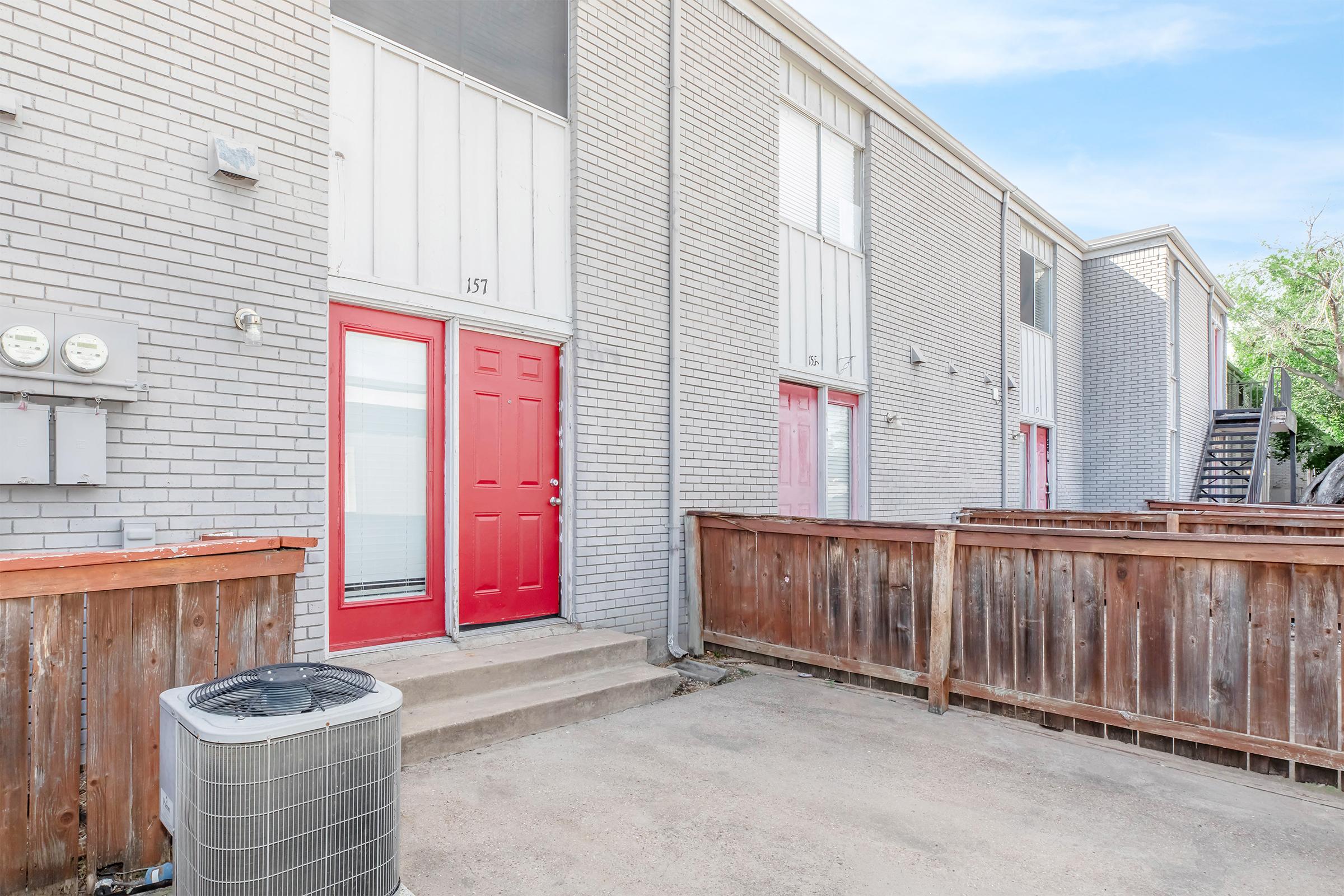 Exterior view of a gray brick apartment complex featuring two units with red doors. A wooden fence surrounds a small outdoor area, and an air conditioning unit is placed on the left side of the image. Visible stairs lead to additional units in the background.