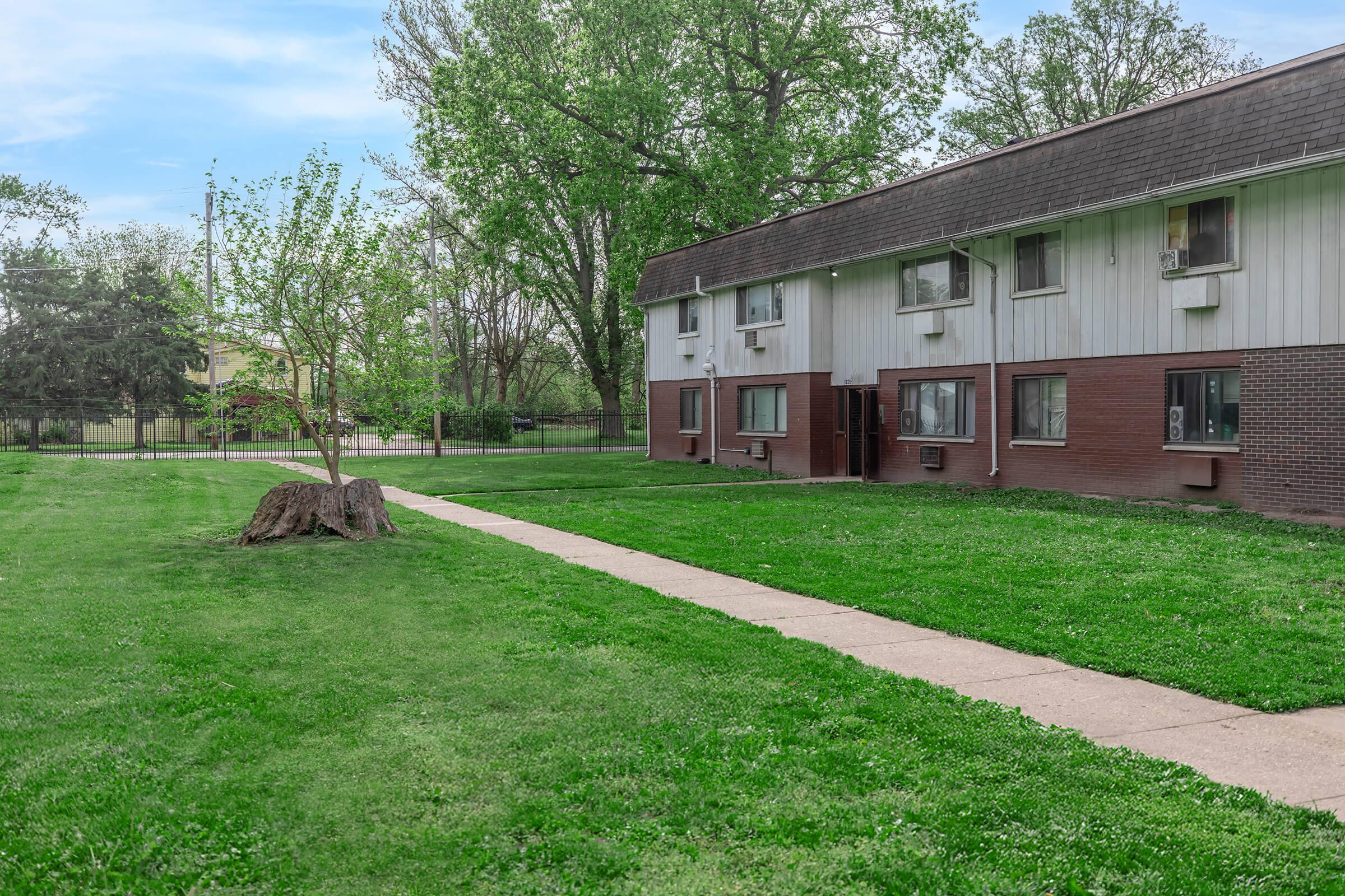 A two-story apartment building with multiple air conditioning units, surrounded by green grass and trees. A concrete walkway leads from the foreground to the building. A large tree stump is visible on the lawn, with clear blue skies in the background.