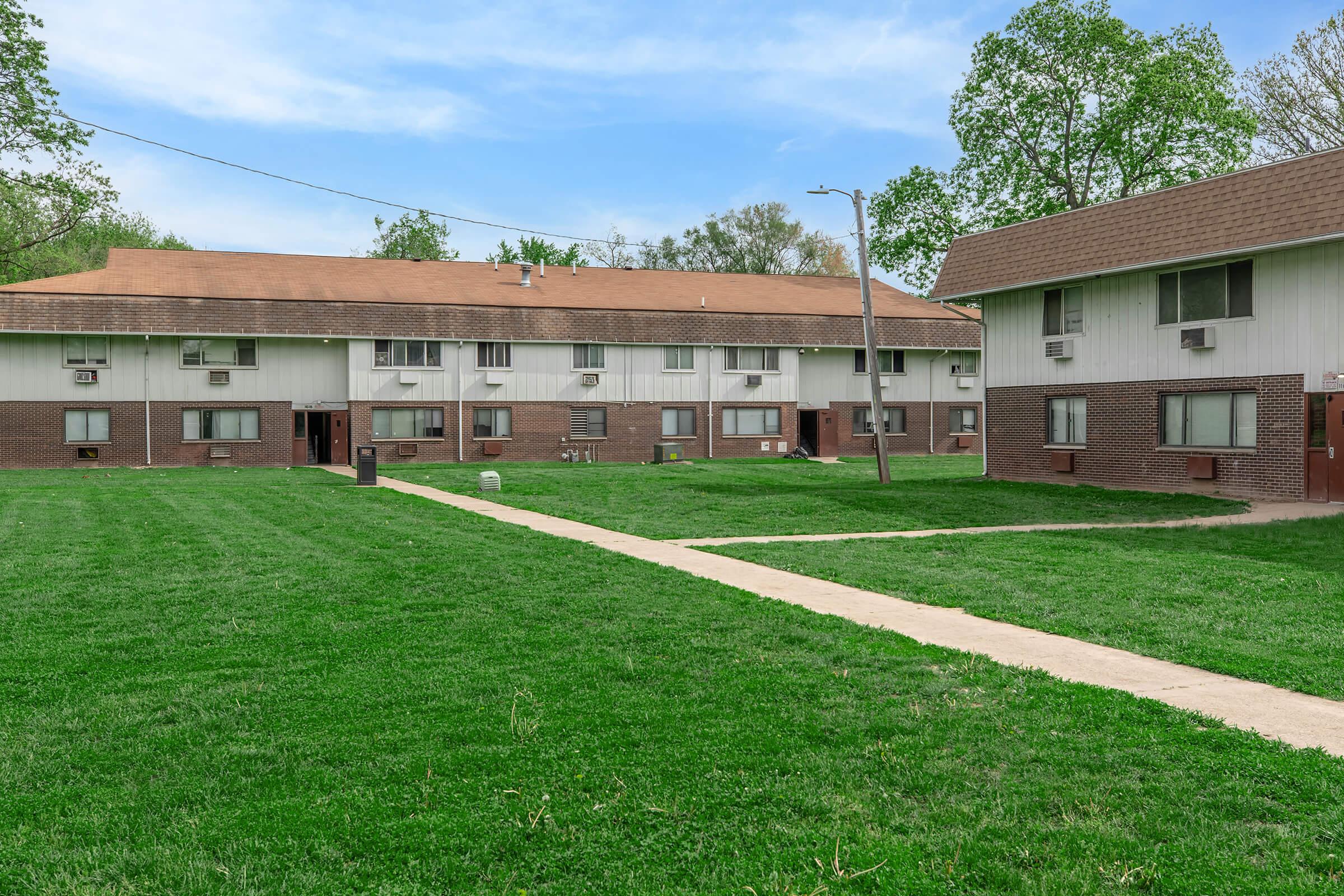 A view of two low-rise apartment buildings with brown and white exteriors, set in a grassy area. The buildings are surrounded by trees and feature multiple windows and air conditioning units. A concrete pathway runs between the buildings, leading through the lawn area.