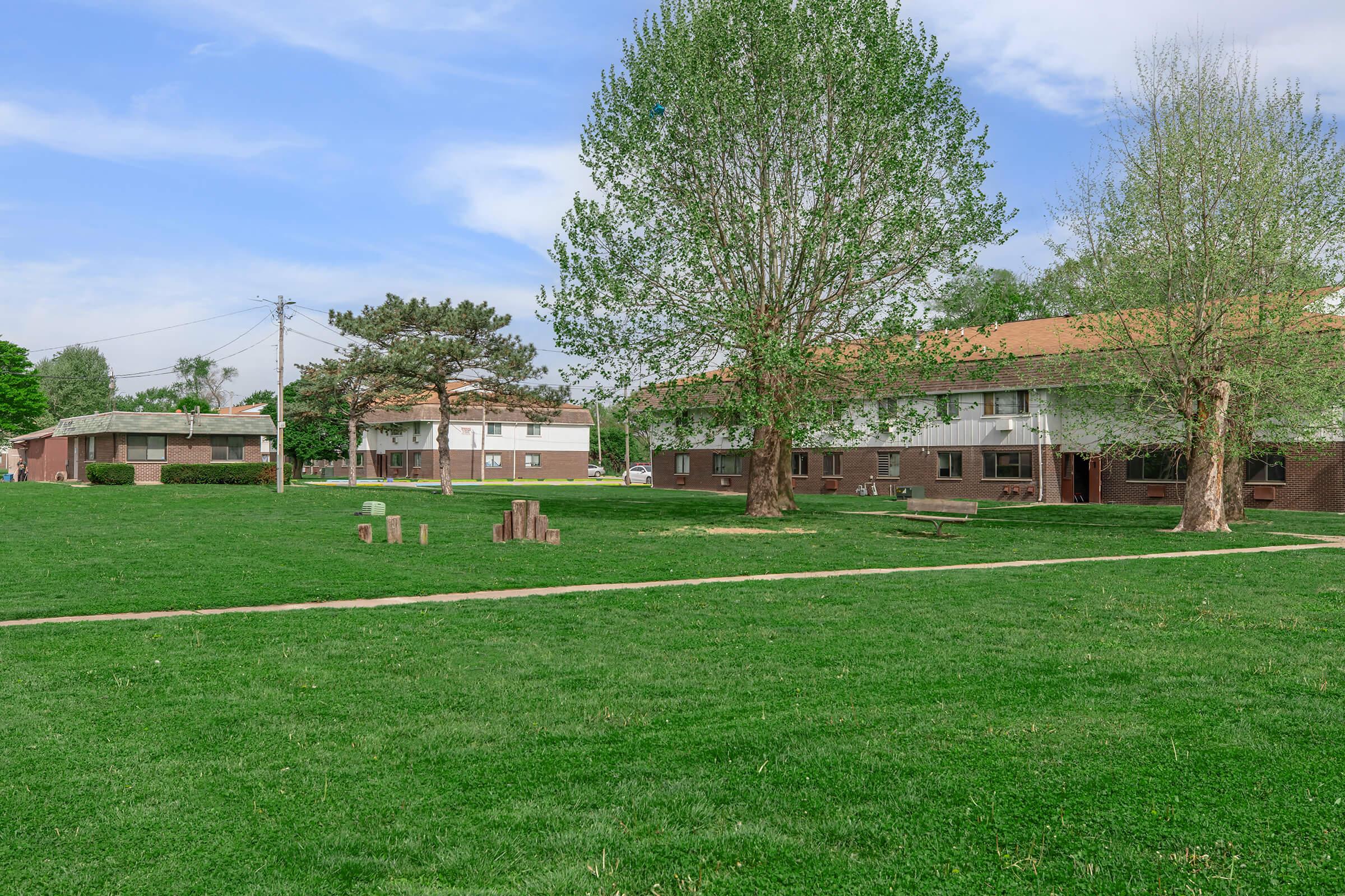 A green lawn with pathways and trees, surrounded by several low-rise buildings. Some furniture is placed on the lawn, and power lines are visible in the background under a clear blue sky.