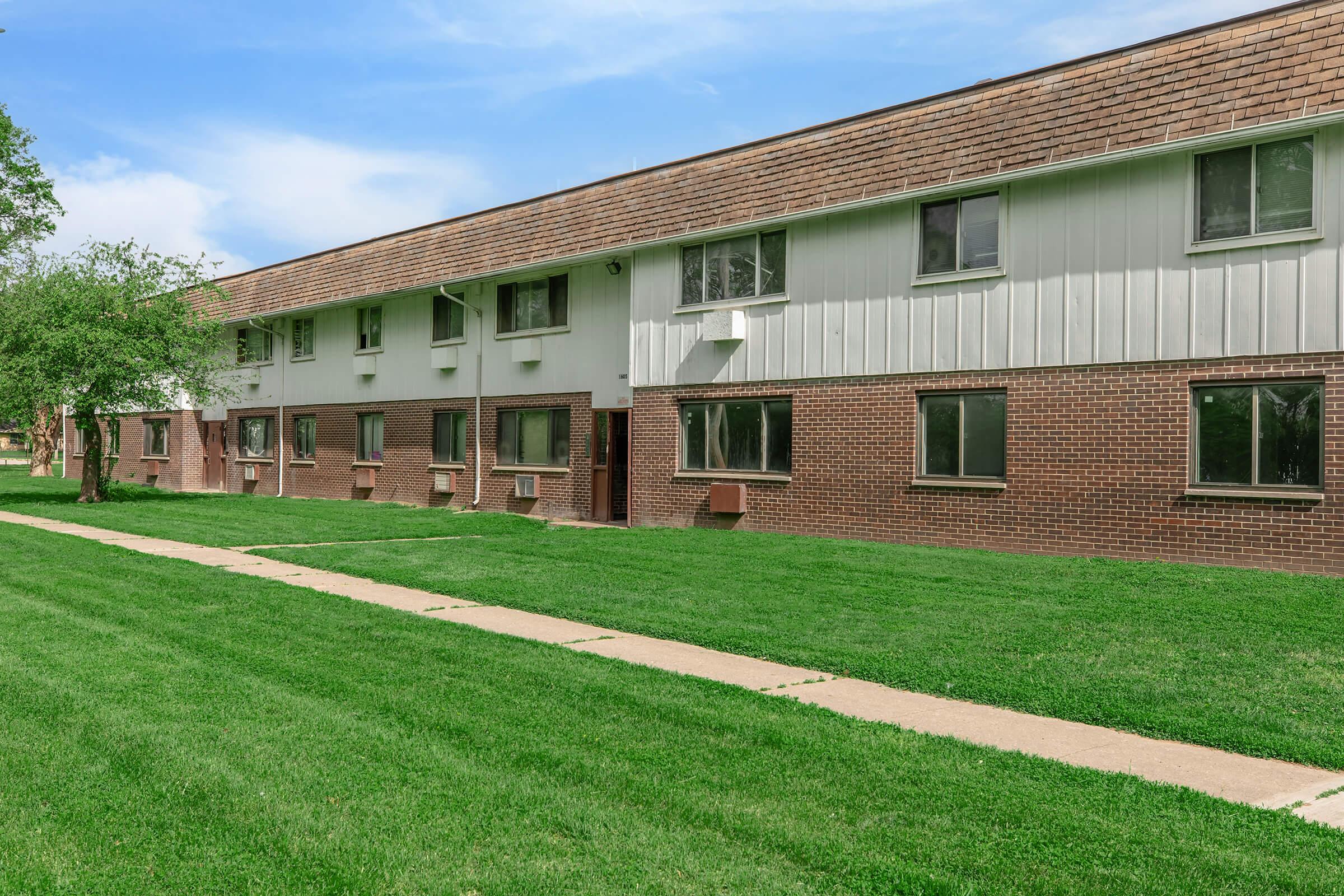 A low-rise apartment building with a brick and white exterior, surrounded by well-maintained green grass. The building features multiple windows and a walkway in front, with some trees visible in the background under a clear blue sky.
