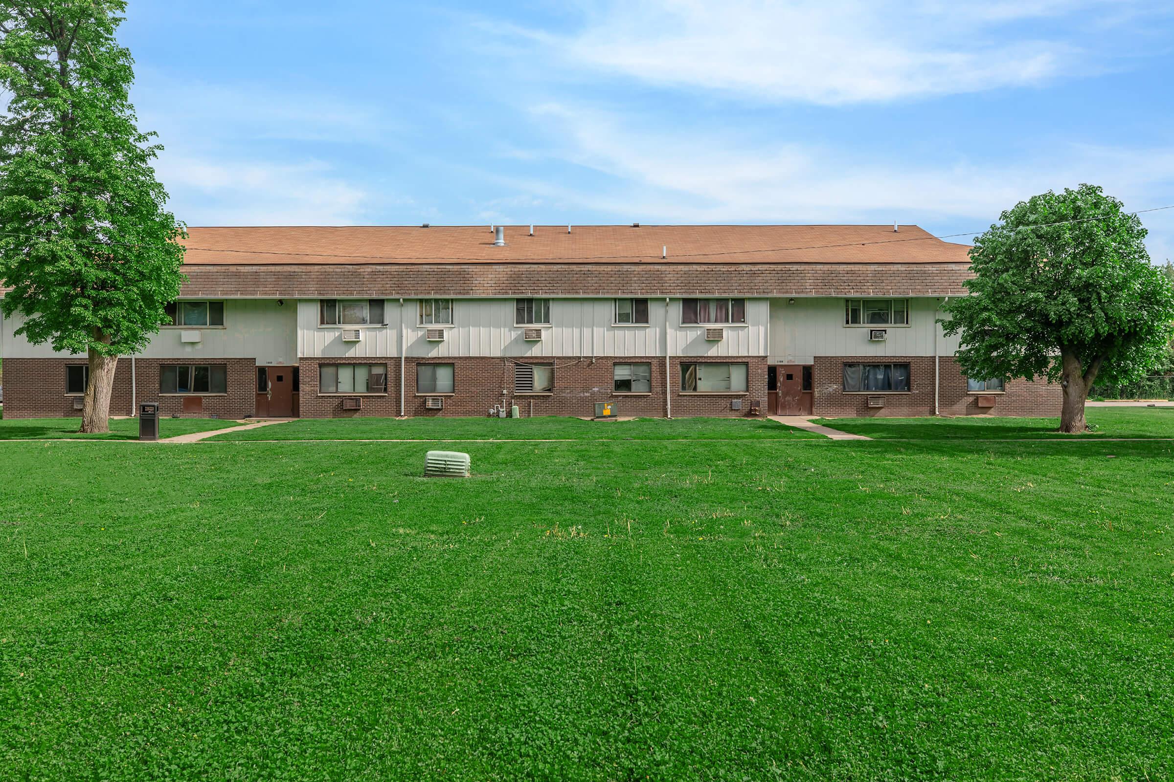A view of a multi-unit residential building with a brown exterior and multiple air conditioning units. The building is surrounded by a well-maintained green lawn, featuring two large trees on either side. The sky is clear with a few clouds.