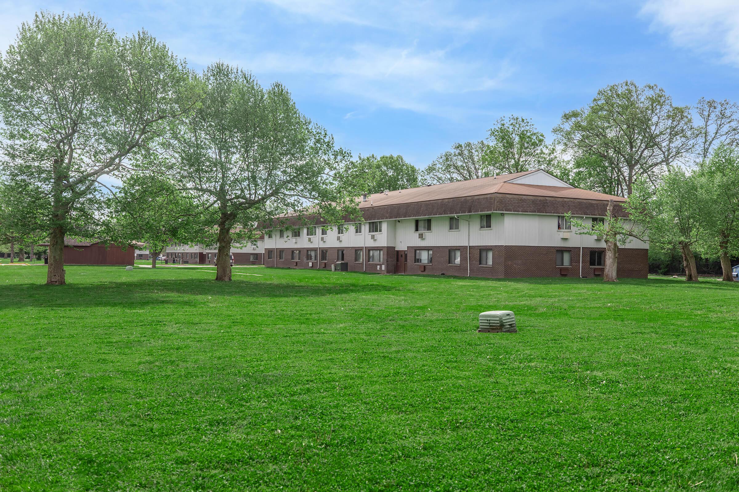 A grassy area with two large trees in the foreground, leading to a multi-story building with a brown and white exterior. The scene features a clear blue sky and a few clouds, creating a peaceful outdoor setting.