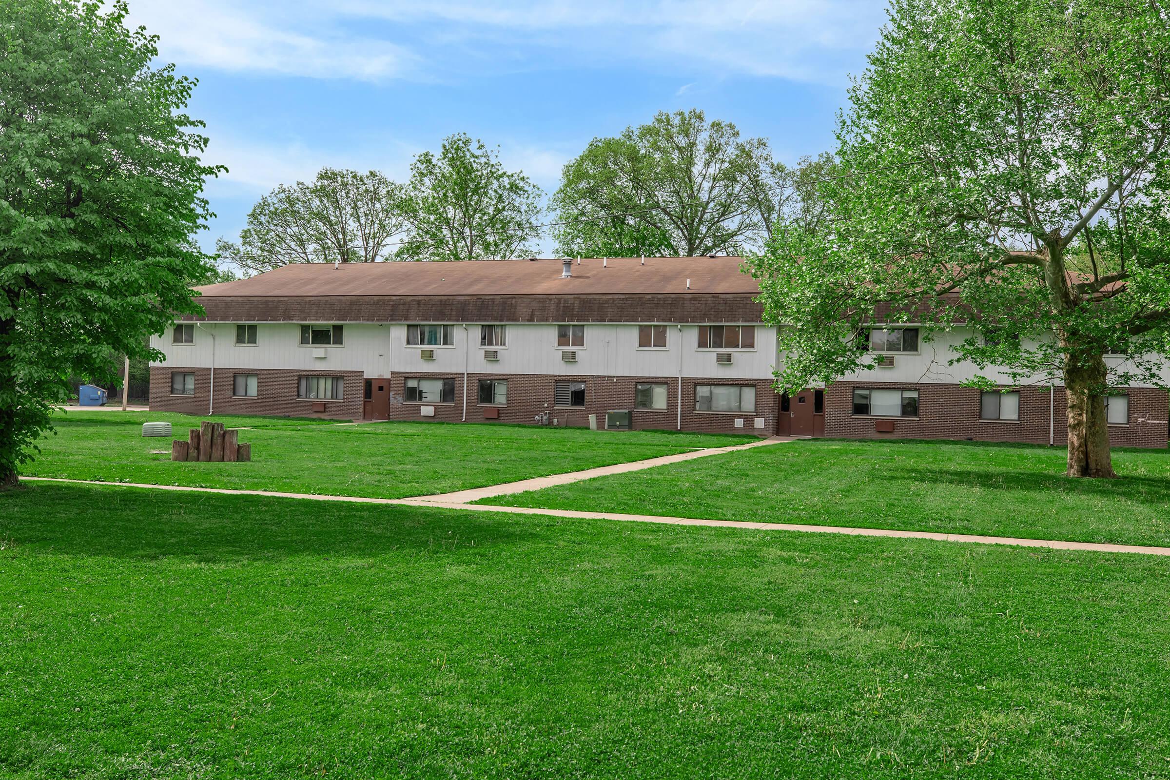 A low-rise apartment building with brown siding, situated in a grassy area. The building features multiple windows and air conditioning units. Surrounding the structure are trees and a neatly maintained lawn. A pathway runs through the grass, leading to the entrance of the building.