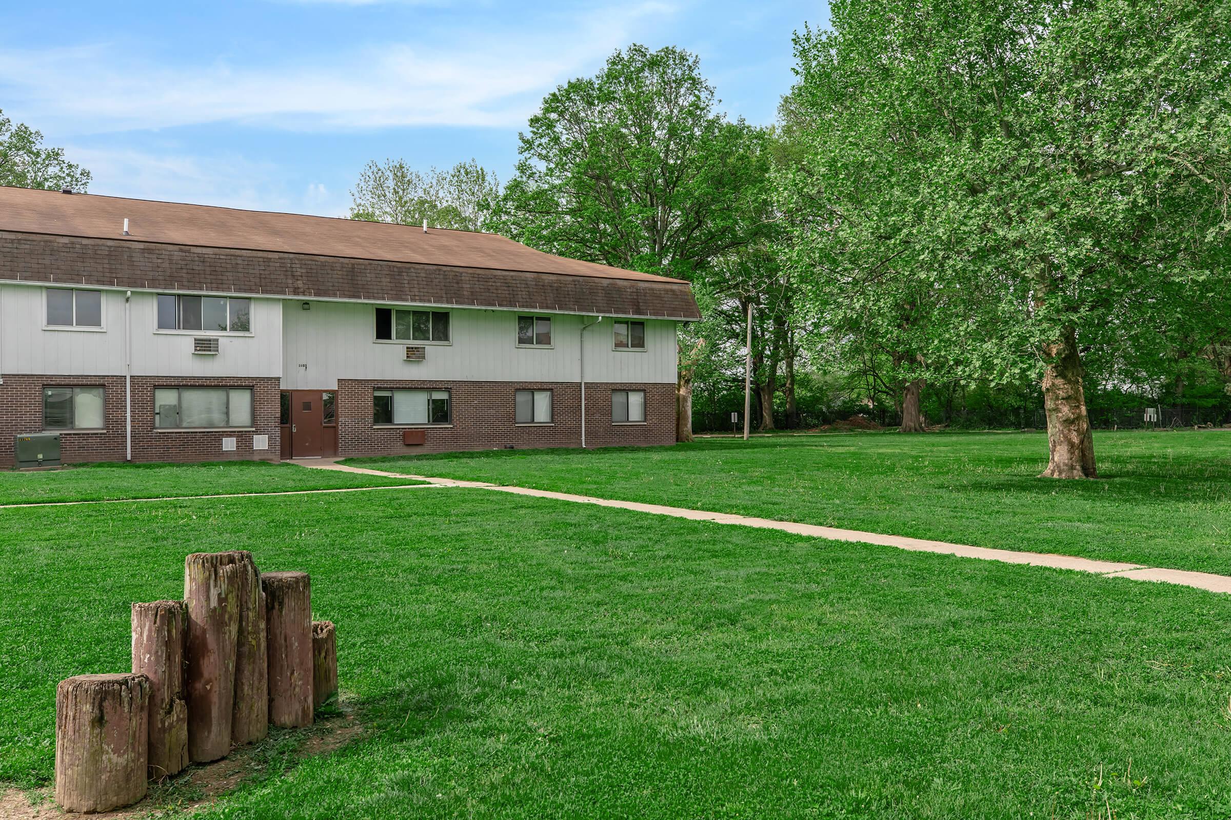 A low-rise apartment building with a brown roof, surrounded by green lawn and trees. A paved pathway leads from the foreground to the entrance of the building. In the foreground, there are wooden stumps arranged in a cluster. The sky is clear with a few clouds, indicating a sunny day.