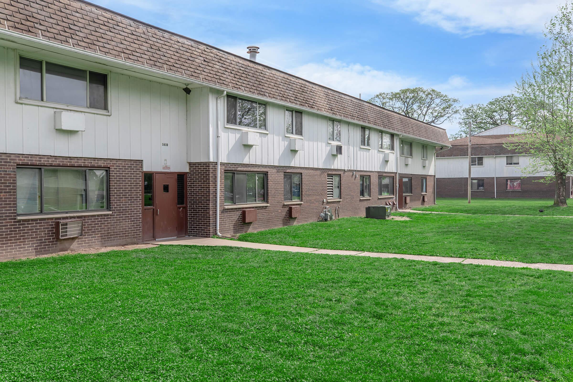 A two-story residential building with white siding and brown brick accents. The exterior features several windows and a single front door. Green grass surrounds the building, and another similar structure is visible in the background under a clear blue sky.