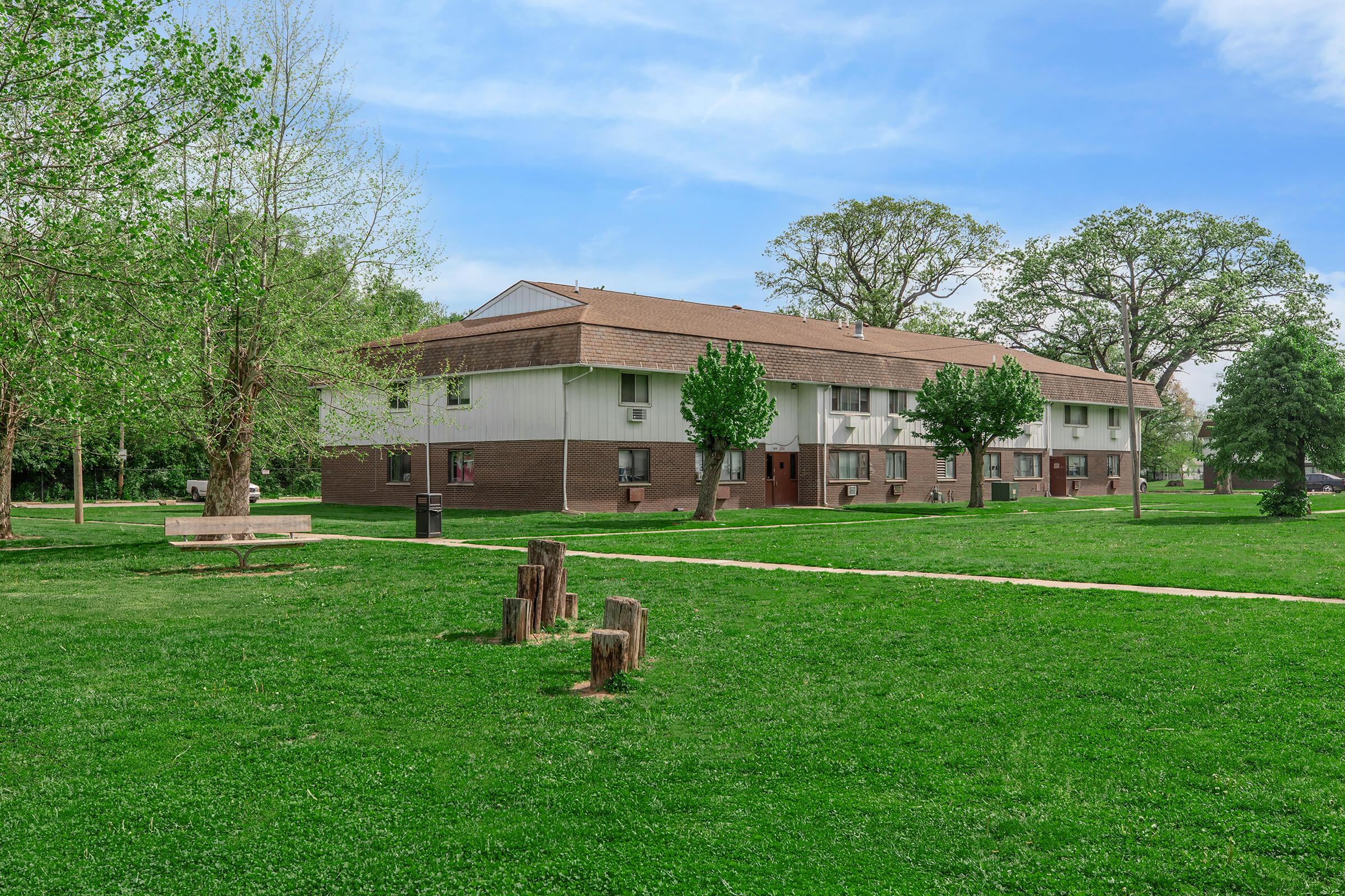 A two-story apartment building situated in a green park area, surrounded by trees. In the foreground, there is a grassy lawn with wooden stumps and a walking path. Bright blue sky with scattered clouds adds to the serene atmosphere of the setting.