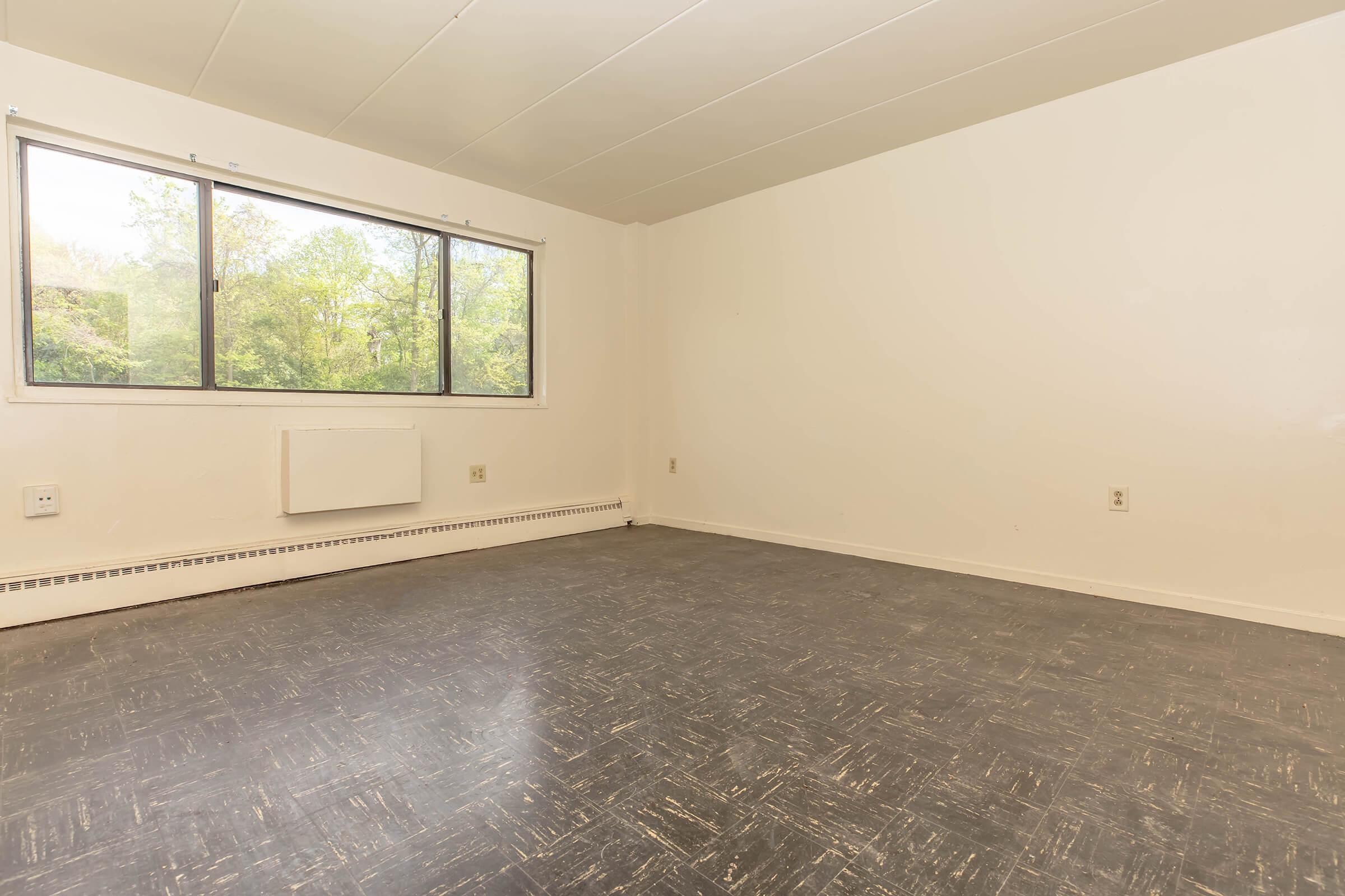 A spacious, empty room with light-colored walls and a large window offering a view of greenery outside. The floor is dark with a textured pattern, and there is a baseboard heater mounted on the wall. The overall atmosphere is bright and open, suggesting potential for personalization and decoration.