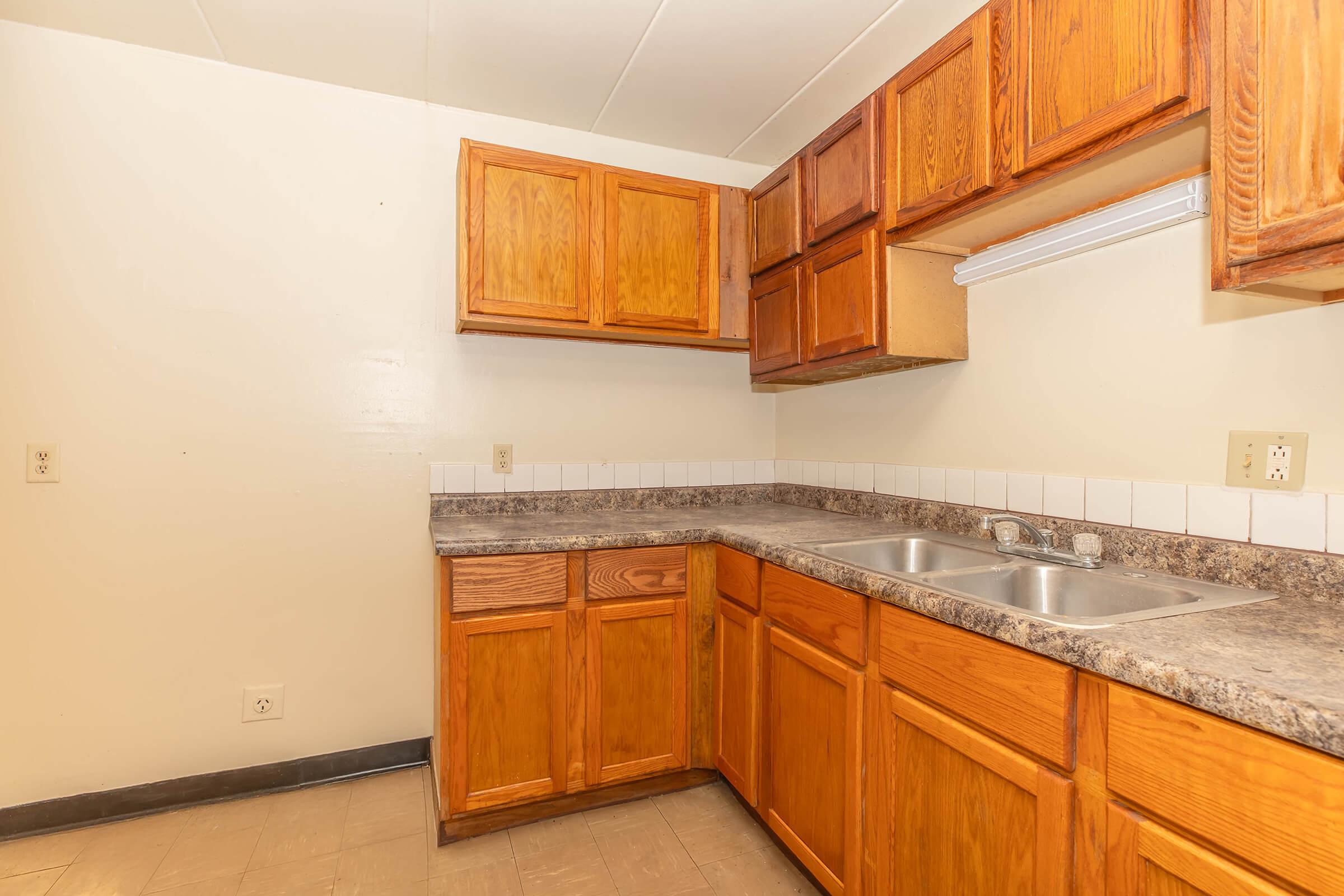 A simple kitchen featuring wooden cabinets, a countertop with a double sink, and beige walls. The cabinets are above and below the countertop, with a white backsplash. The floor is light-colored, and there is a small window sill visible above the sink area.