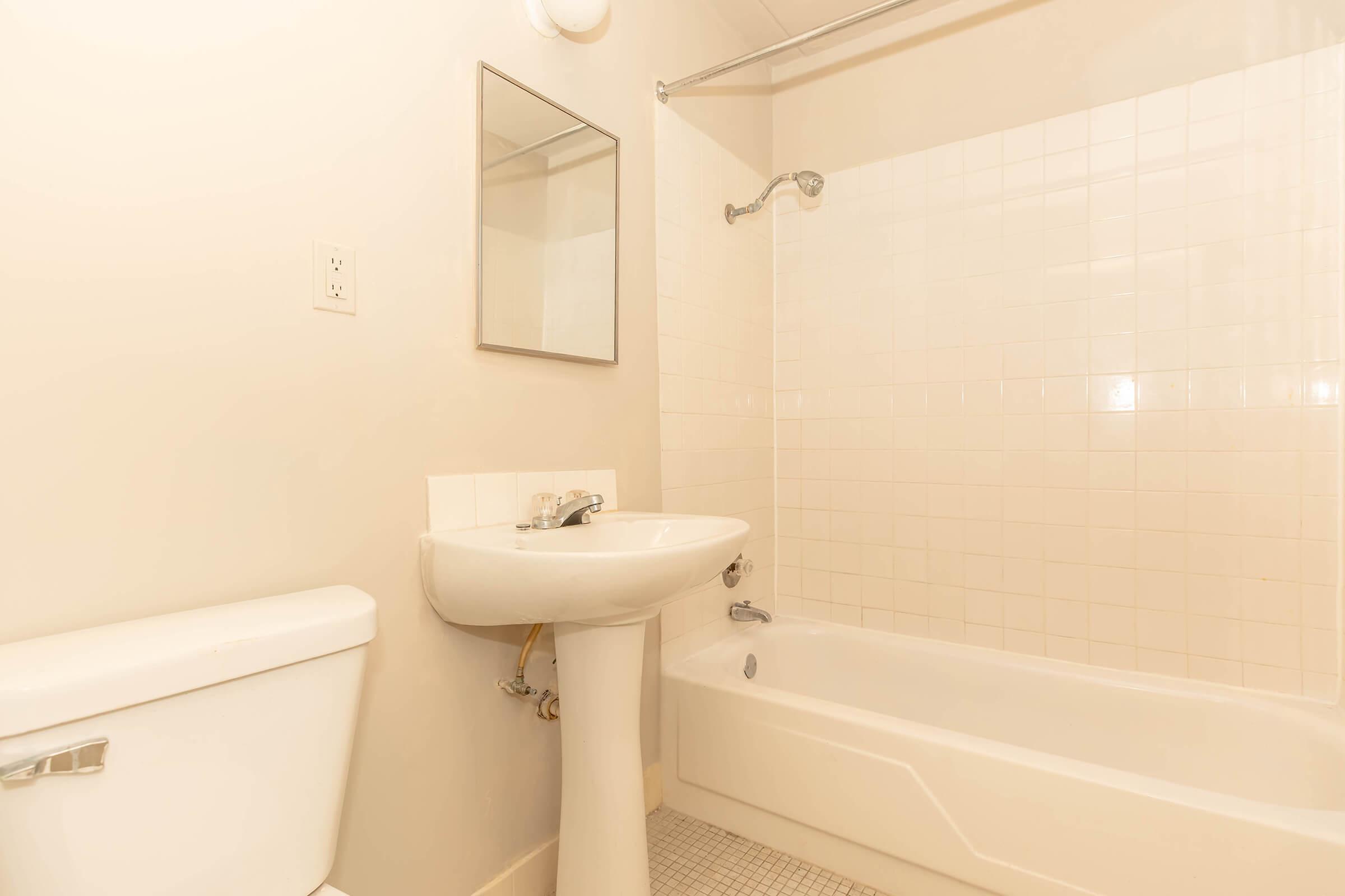 A clean and simple bathroom featuring a bathtub with a shower head, a white pedestal sink, and a toilet. The walls are light-colored, and there is a small mirror above the sink. The floor is tiled in a subtle pattern. The overall design is minimalist and modern.