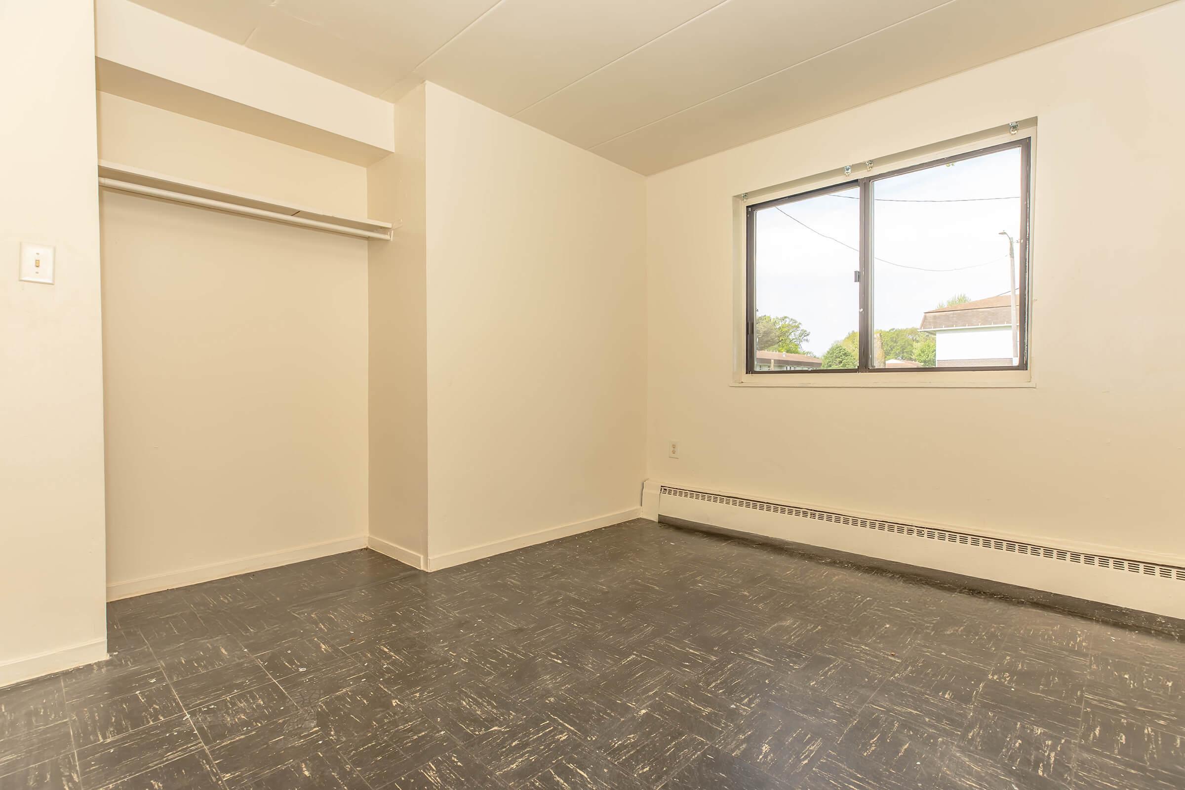 A vacant room with light-colored walls, a window allowing natural light, and a closet without doors. The floor has a dark linoleum pattern, and there are no furnishings or decorations, creating an open and spacious atmosphere.