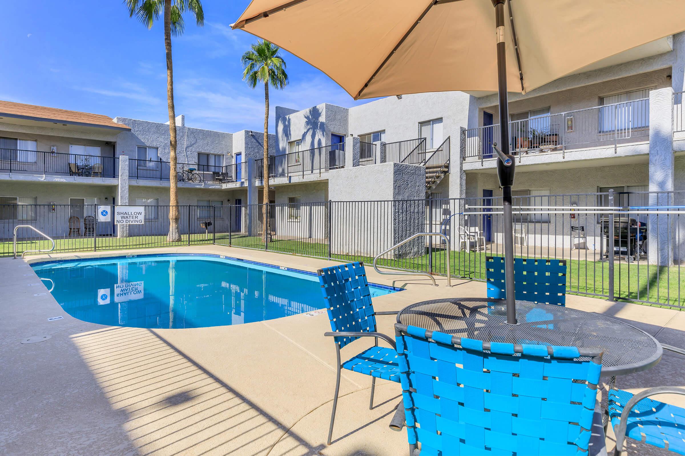 A swimming pool surrounded by palm trees and lounge chairs. The area includes a table with chairs, and there are two-story buildings in the background. The pool is clean and inviting, with a bright blue sky overhead, creating a relaxing atmosphere for residents.