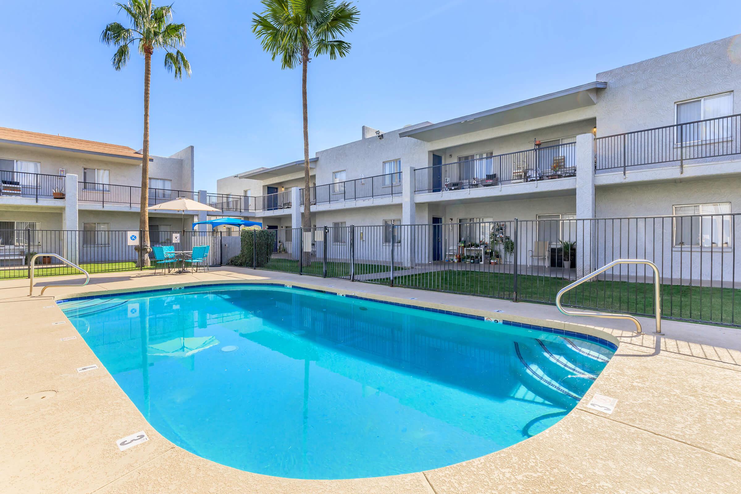 A clear swimming pool surrounded by a fenced patio area, with palm trees nearby. Two-story apartment buildings are visible in the background, featuring balconies with outdoor furniture. Sunlight creates a bright and inviting atmosphere.