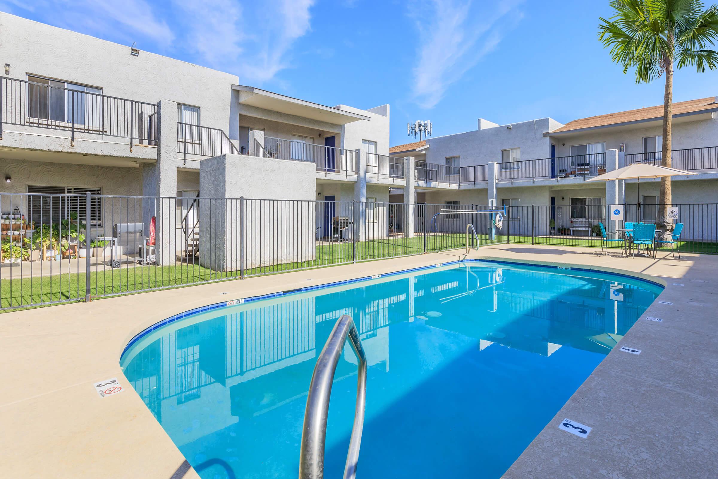 A well-maintained apartment complex featuring a clear swimming pool surrounded by a grassy area. The two-story buildings have balconies and are set against a bright blue sky with a few scattered clouds. Palm trees add a tropical touch to the scene, creating a relaxed atmosphere.