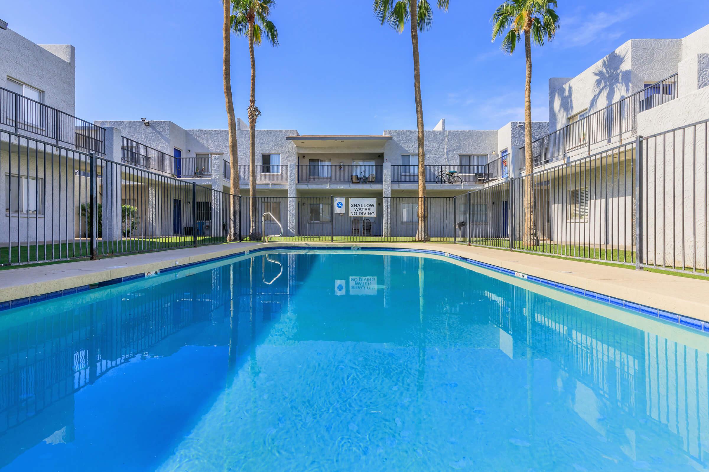 A clear, inviting swimming pool surrounded by a gated area, with palm trees lining the background. The pool reflects the blue sky, and there are two-story apartment buildings in the backdrop. The scene is bright and sunny, creating a relaxed atmosphere.