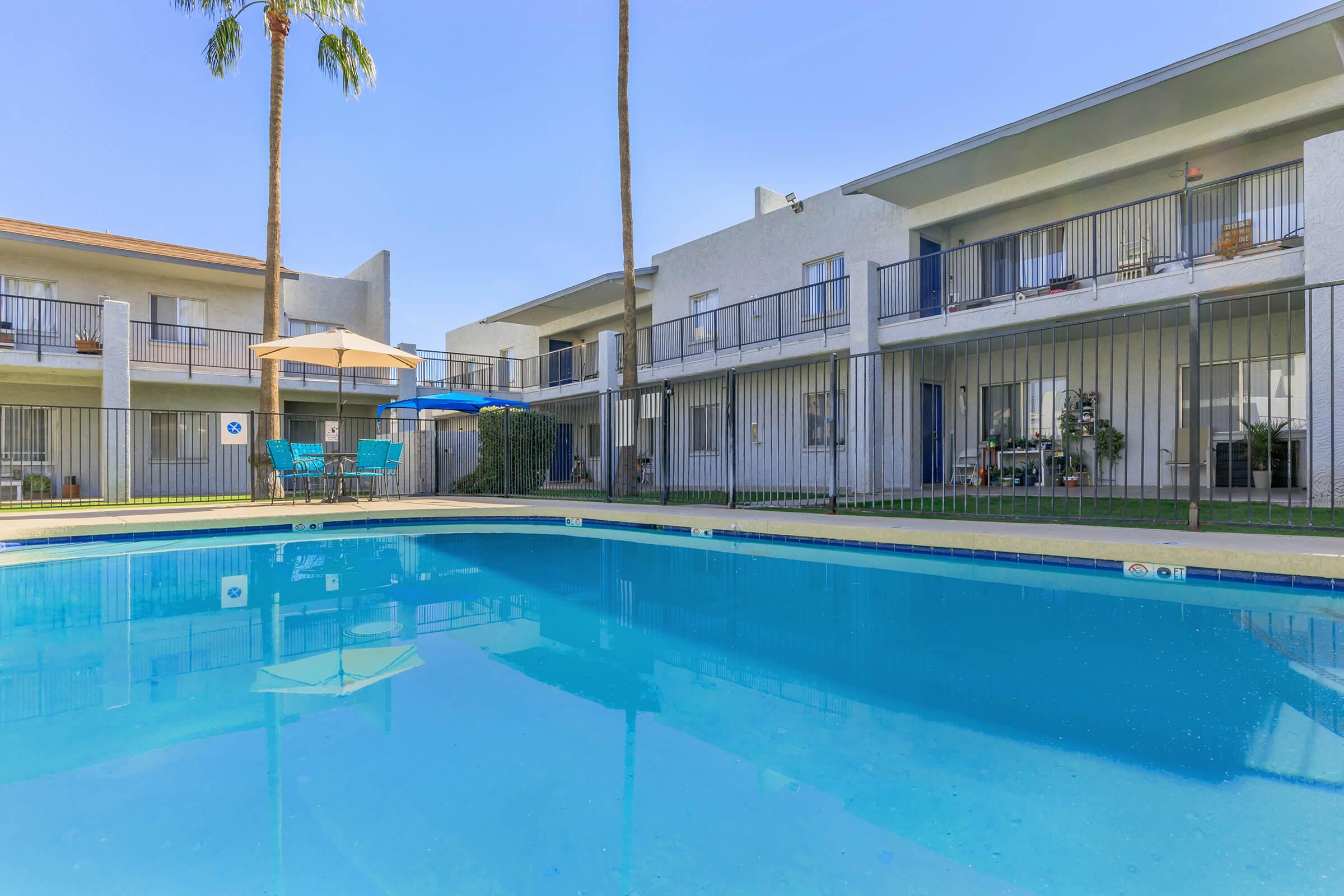 A clear blue swimming pool in the foreground with a fenced area. In the background, there are two-story apartment buildings with balconies. Palm trees are on the side, and patio furniture, including chairs and an umbrella, is visible near the pool. The sky is blue and clear, creating a sunny atmosphere.