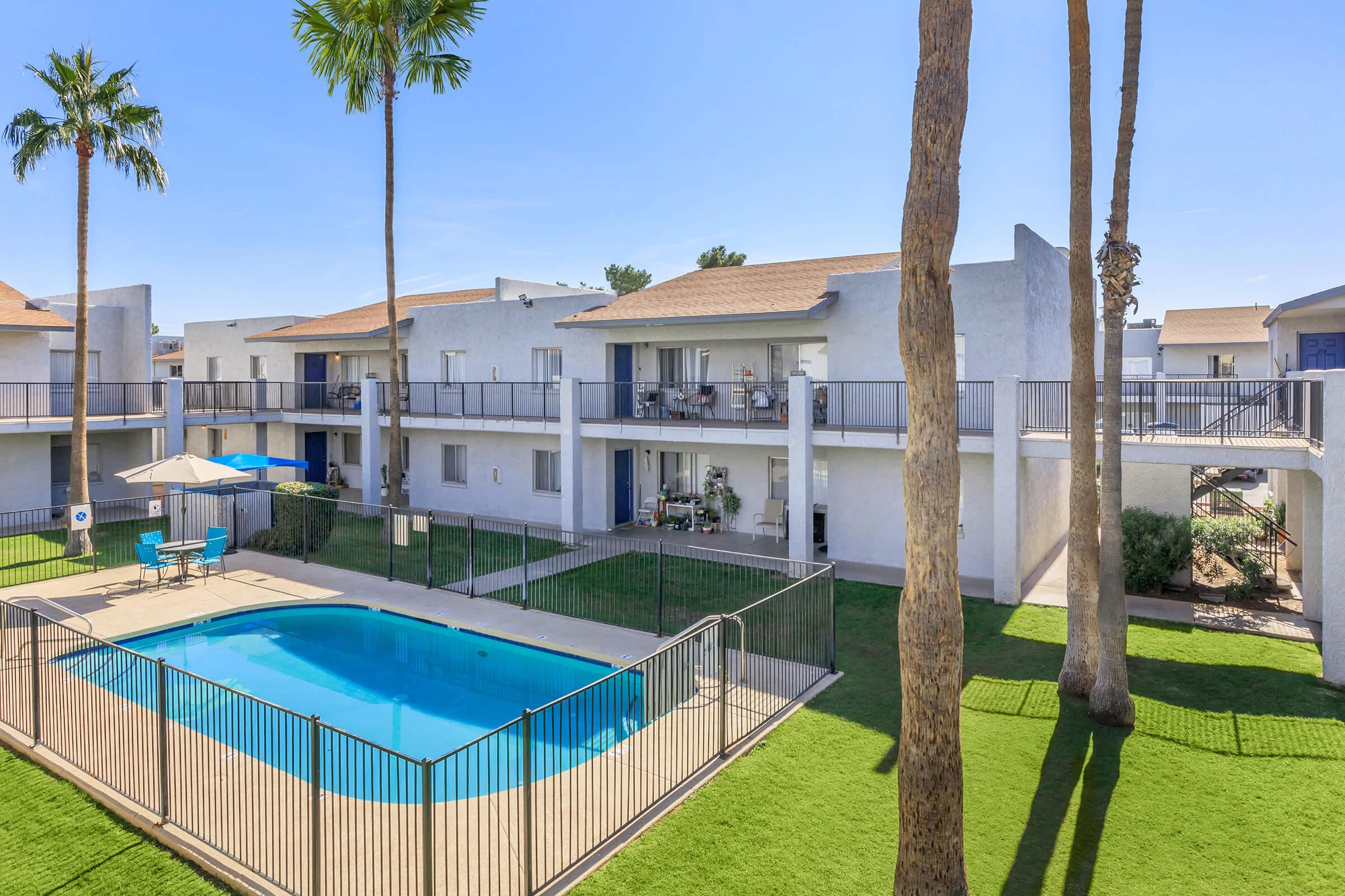 A view of a serene courtyard featuring a swimming pool surrounded by a gated area. Two palm trees stand tall next to the pool. Nearby, two-story residential buildings with balconies are visible, showcasing a clear blue sky and well-manicured green grass.