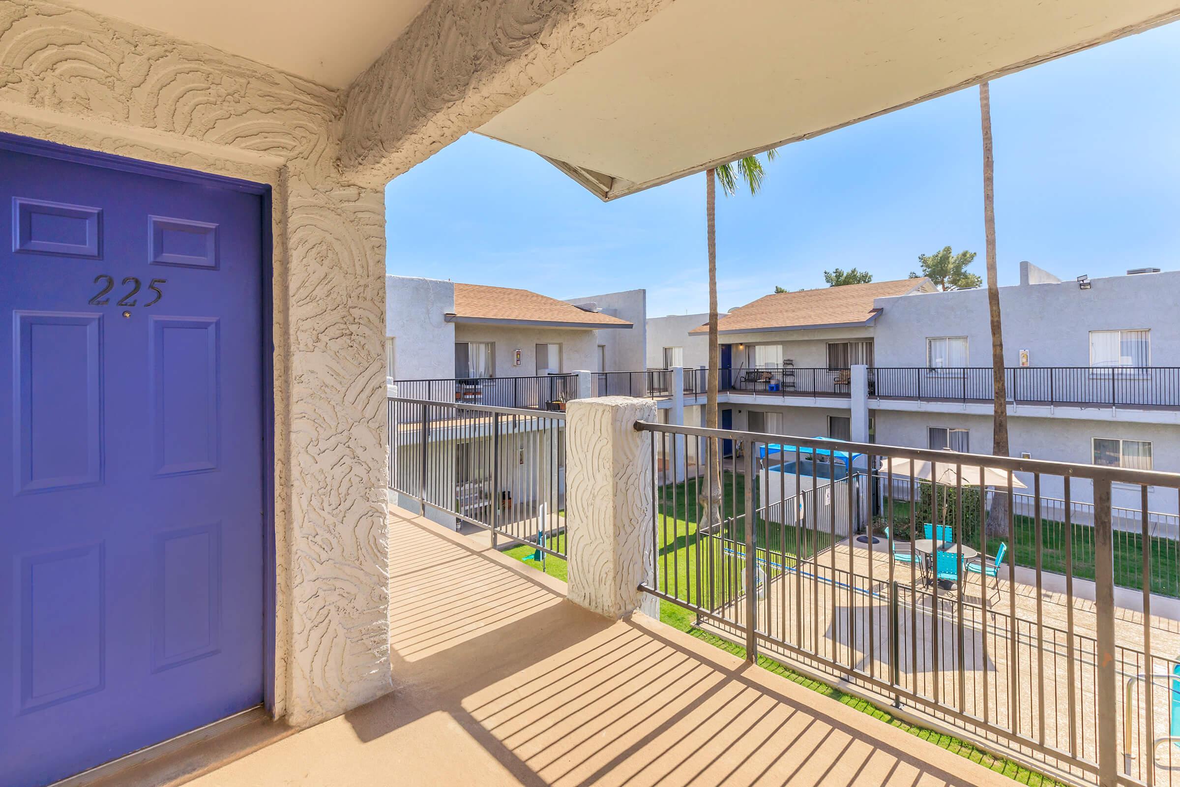 View from a balcony with a bright purple door marked "225." The scene shows a sunny courtyard with grass and lounge chairs surrounded by beige apartment buildings. Palm trees are visible, adding a tropical feel to the setting. The atmosphere is calm and inviting.