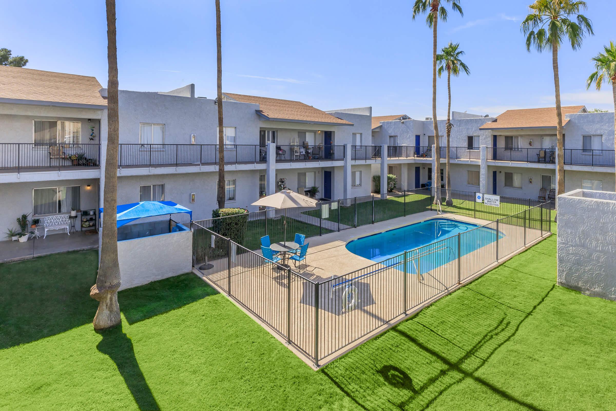 Aerial view of an apartment complex featuring a swimming pool surrounded by a fenced area, with lounge chairs and umbrellas. Palm trees are visible, and the buildings have balconies. The area is landscaped with green grass, creating a relaxed outdoor atmosphere.