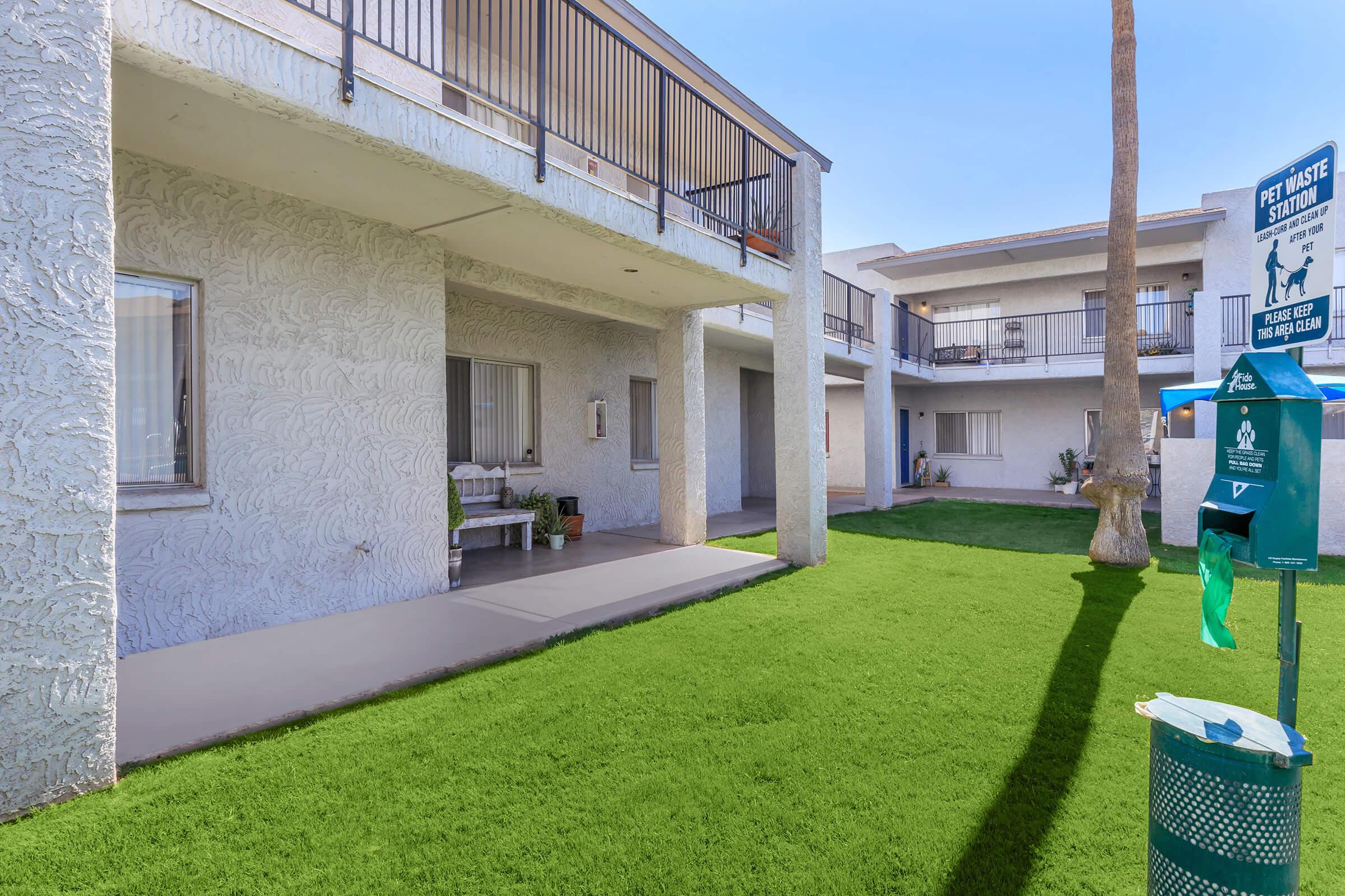 A view of a well-maintained apartment complex featuring two buildings with balconies, surrounded by lush green grass. A pet waste station is visible nearby, and there are potted plants by the entrance of one building, providing a welcoming atmosphere. The sky is clear and blue.