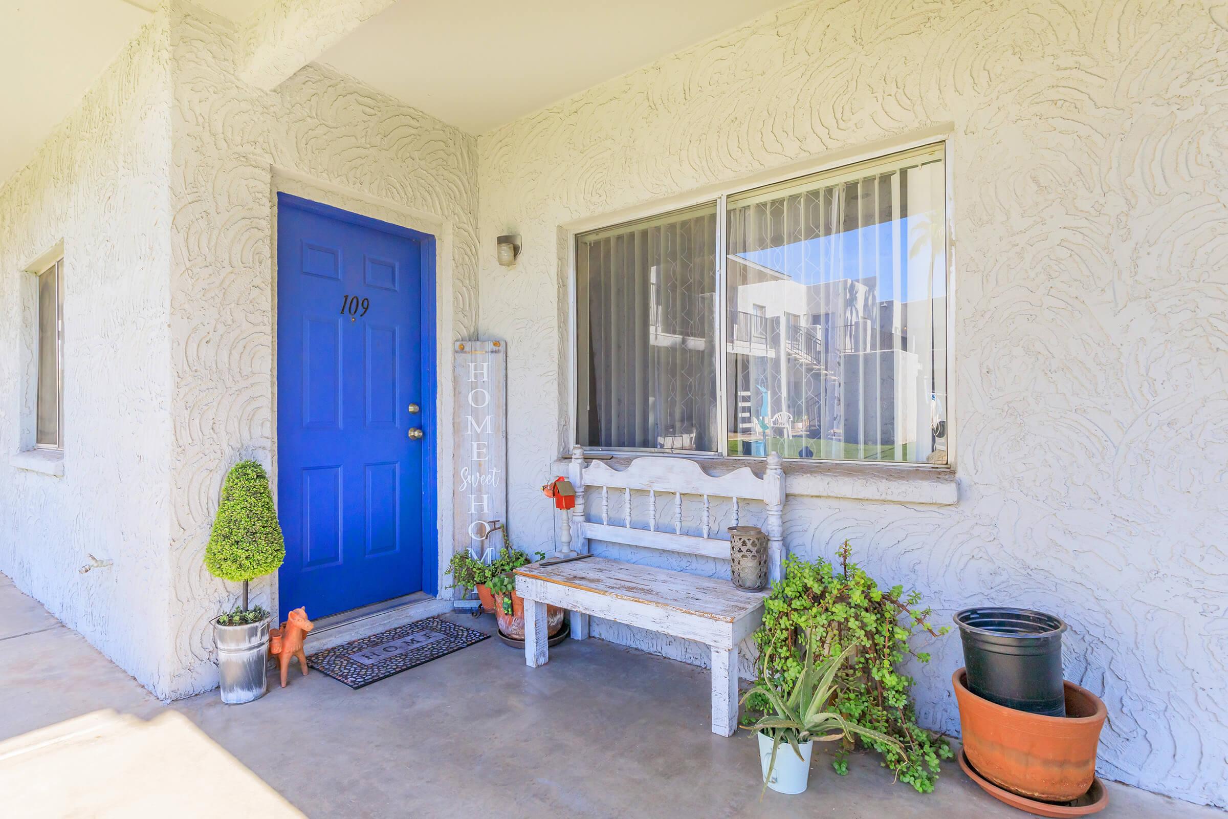 A cozy entrance with a bright blue door labeled '109', featuring a small wooden bench, potted plants, and decorative elements. There is a welcome mat at the entrance, creating an inviting atmosphere. Sunlight casts a warm glow on the textured, light-colored wall.