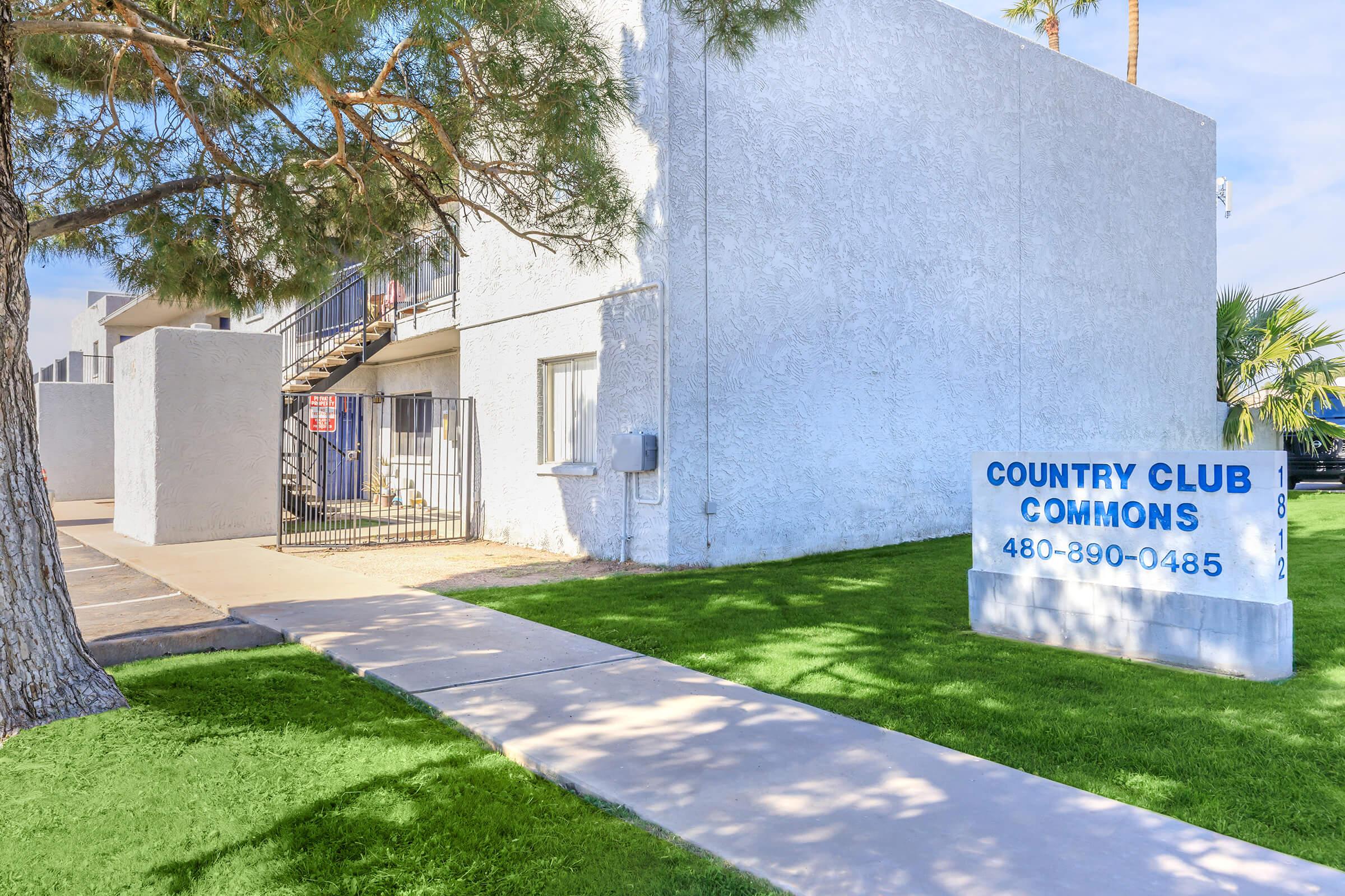 Exterior view of Country Club Commons, featuring a grey building with a sign displaying the name and contact number. The entrance is surrounded by green grass and a tree. Staircase access to upper levels can be seen on the left.