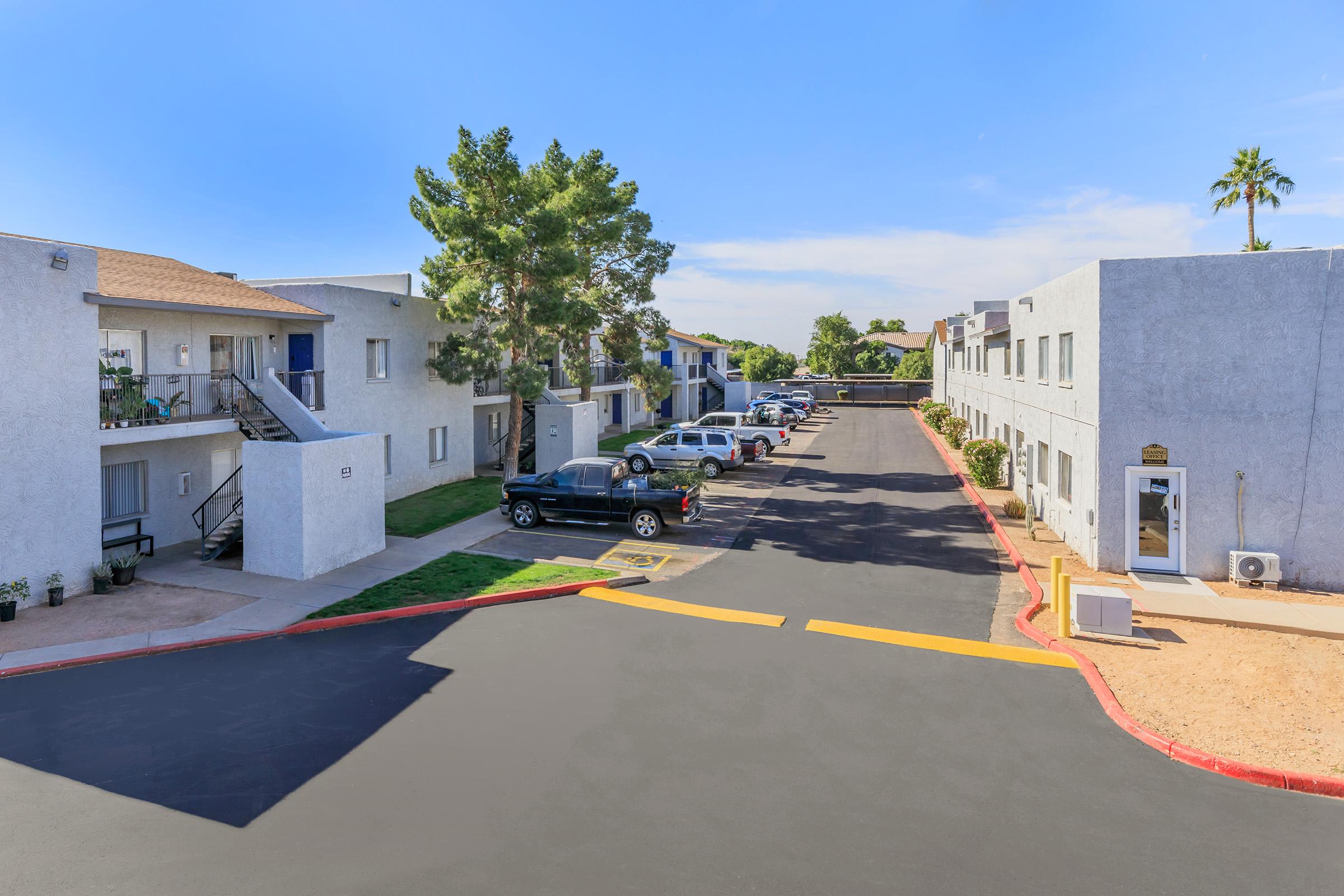 View of a residential complex featuring two-story buildings with a light gray exterior. A parking lot is visible, with several parked cars lined up along the asphalt road. Green trees and shrubs are present, adding to the outdoor scenery, under a clear blue sky.