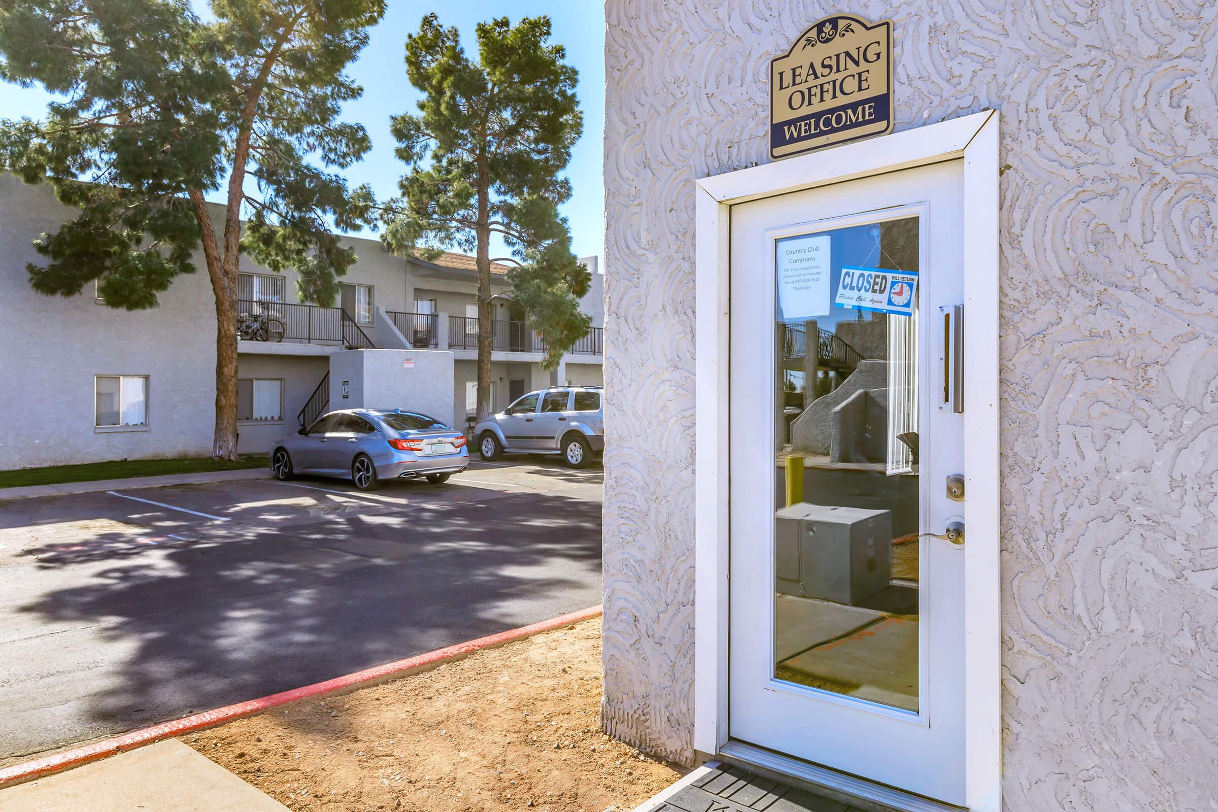 A leasing office entrance with a "Welcome" sign. The glass door features a "Closed" notice. In the background, two parked cars are visible, and the surrounding area includes a few trees and an apartment building. The scene is sunny and well-lit.