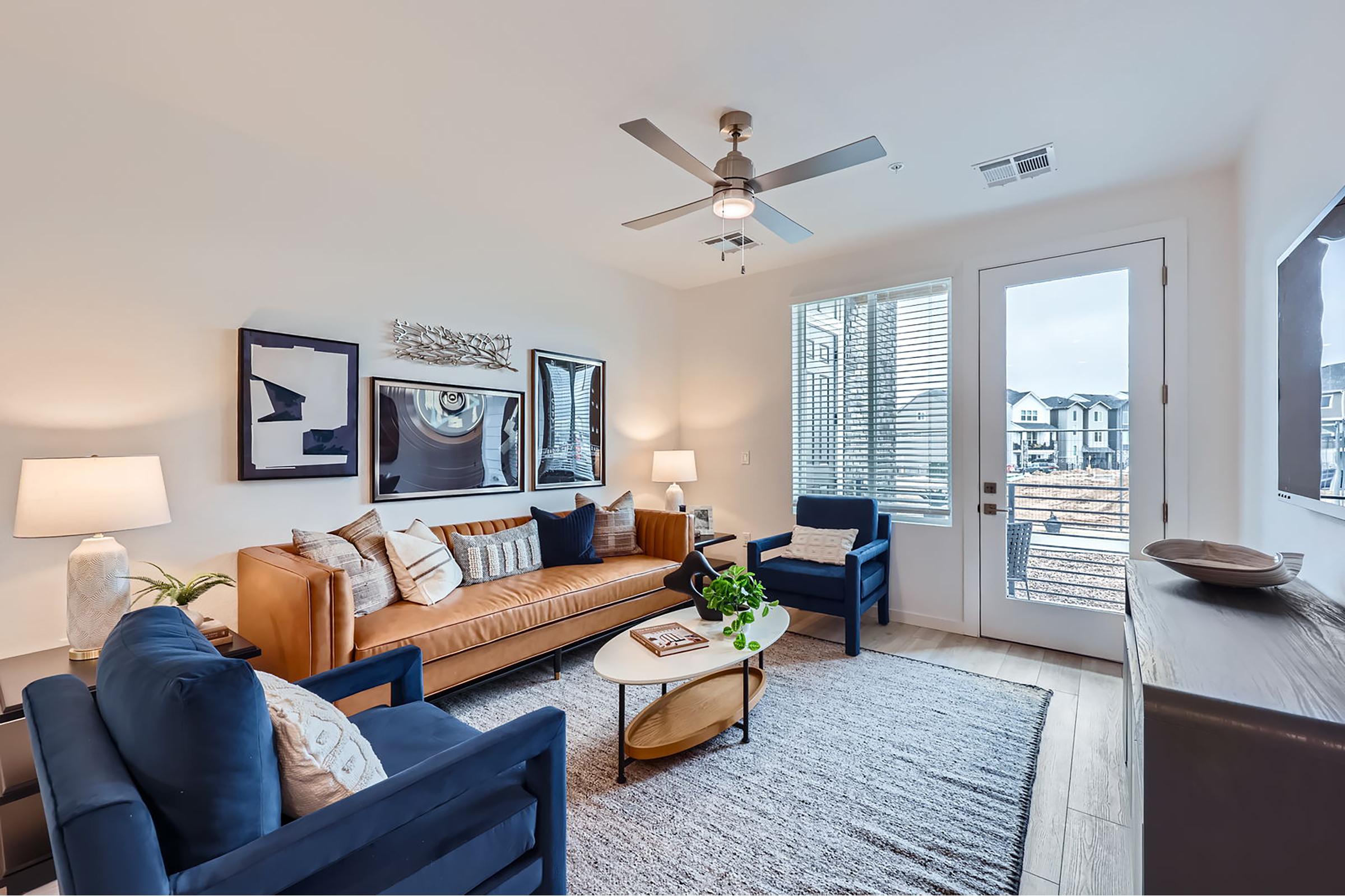 A modern living room featuring a tan leather sofa, two blue armchairs, and a round coffee table on a textured rug. There are artworks on the walls, a lamp providing soft light, and a large window, allowing natural light to fill the space. The decor is sleek and minimalist, with a cozy ambiance.