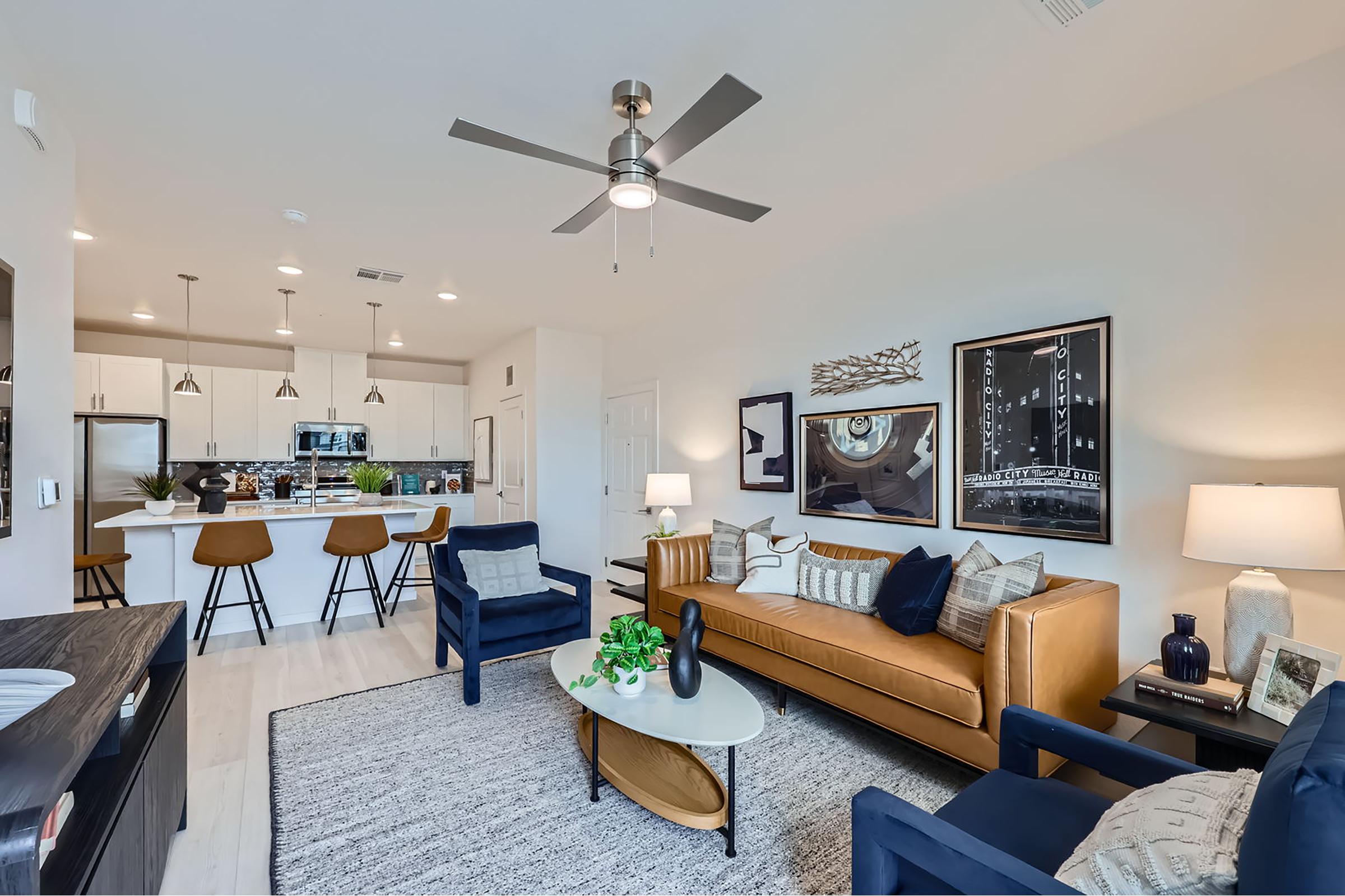 A modern living room featuring a stylish brown couch, two dark blue chairs, and a coffee table with decorative plants. The space includes a ceiling fan, light-colored walls, and artwork. In the background, there is an open kitchen with bar stools and contemporary appliances, creating a warm and inviting atmosphere.