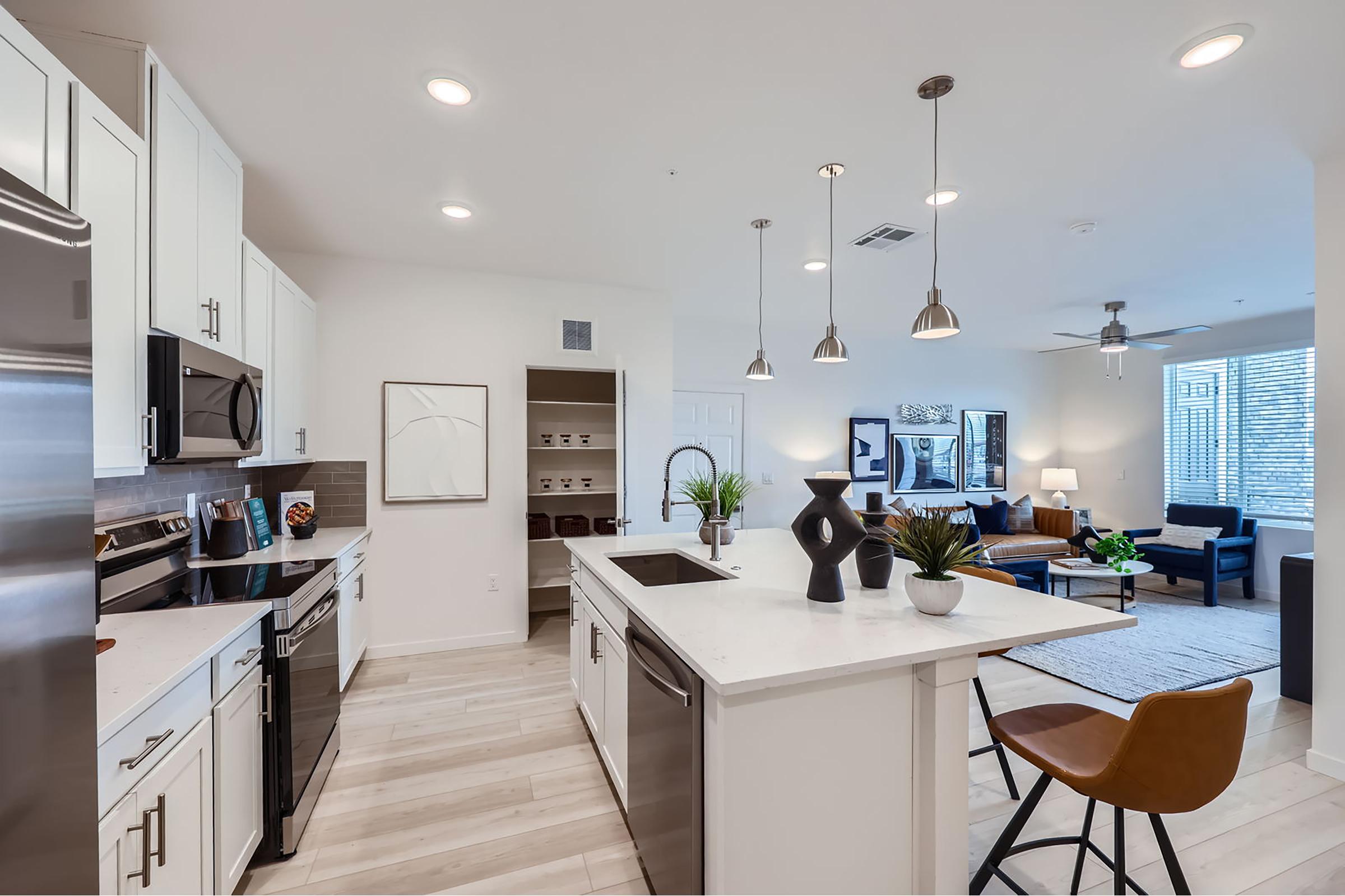 Modern kitchen with white cabinetry, stainless steel appliances, and a large island. Stylish pendant lights hang above the island, which is set with decorative items. The open space leads into a living area with a couch, chairs, and natural light from large windows, creating a bright and inviting atmosphere.