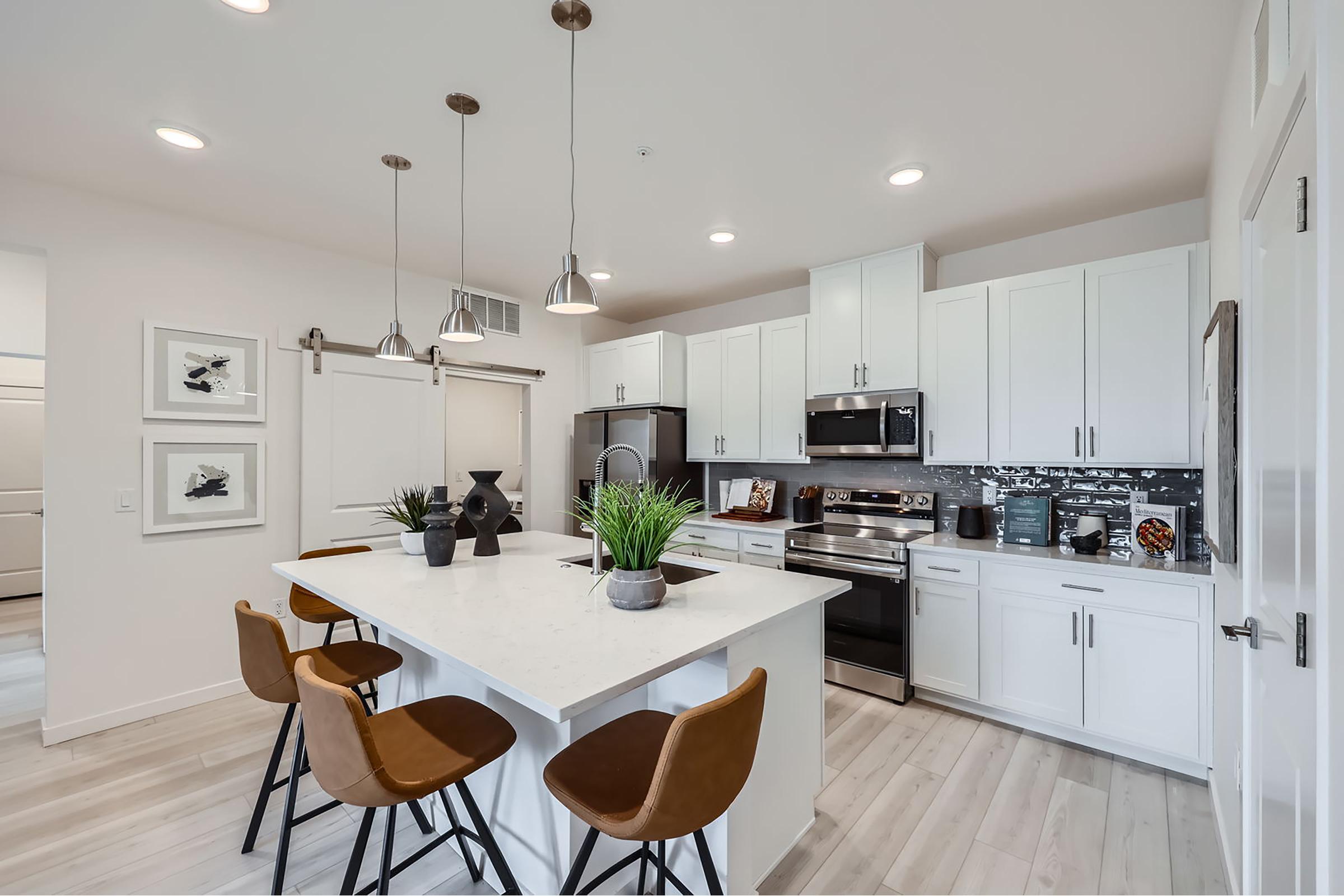 Modern kitchen featuring white cabinetry, stainless steel appliances, and a large island with seating. The space is well-lit with pendant lights and has a clean, contemporary design. Potted plants add a touch of greenery, enhancing the inviting atmosphere of the room.
