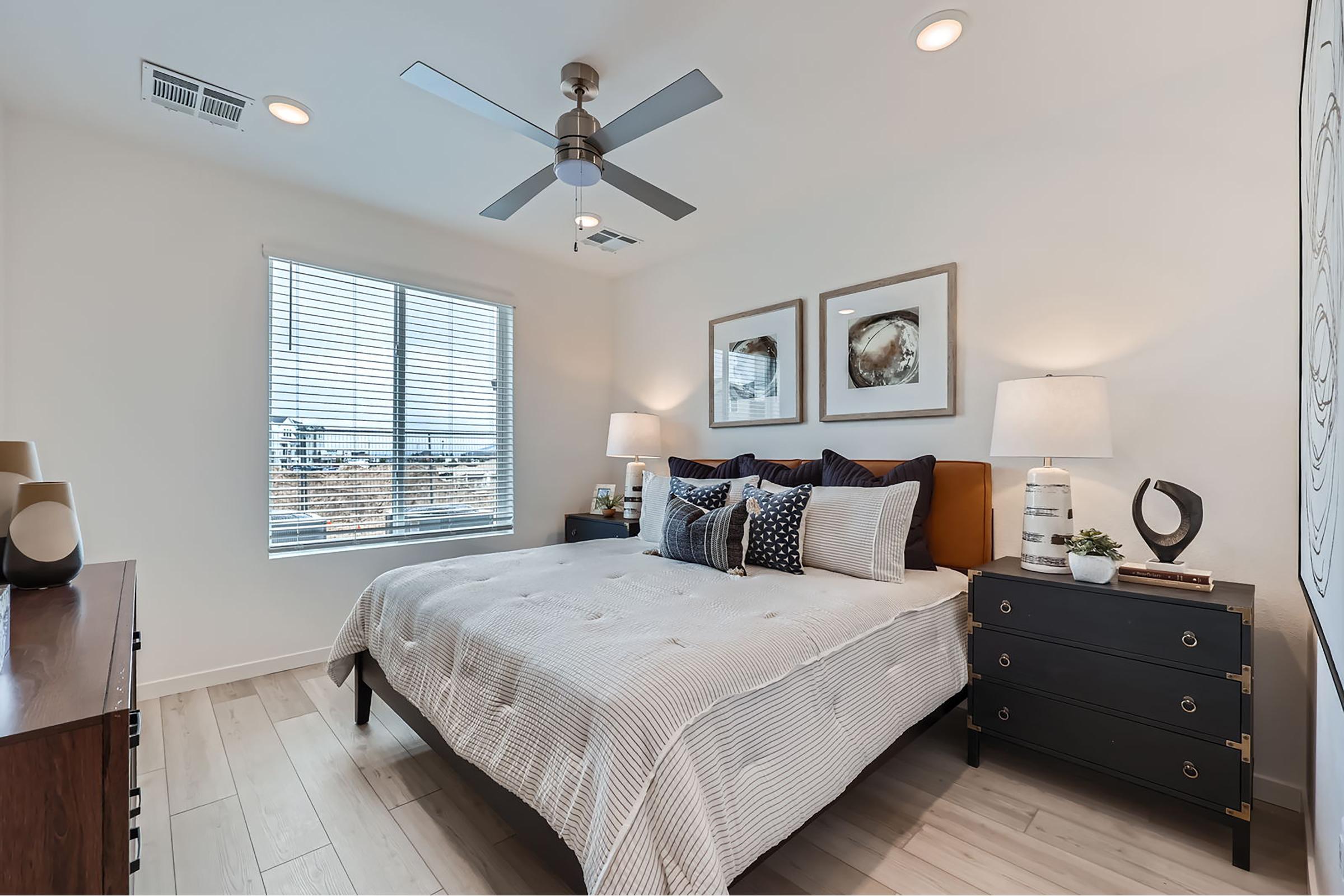 A modern bedroom featuring a neatly made bed with decorative pillows, a wooden dresser, and bedside tables with lamps. There is a ceiling fan above and large windows with blinds, allowing natural light to brighten the space. Two framed artworks are mounted on the wall.