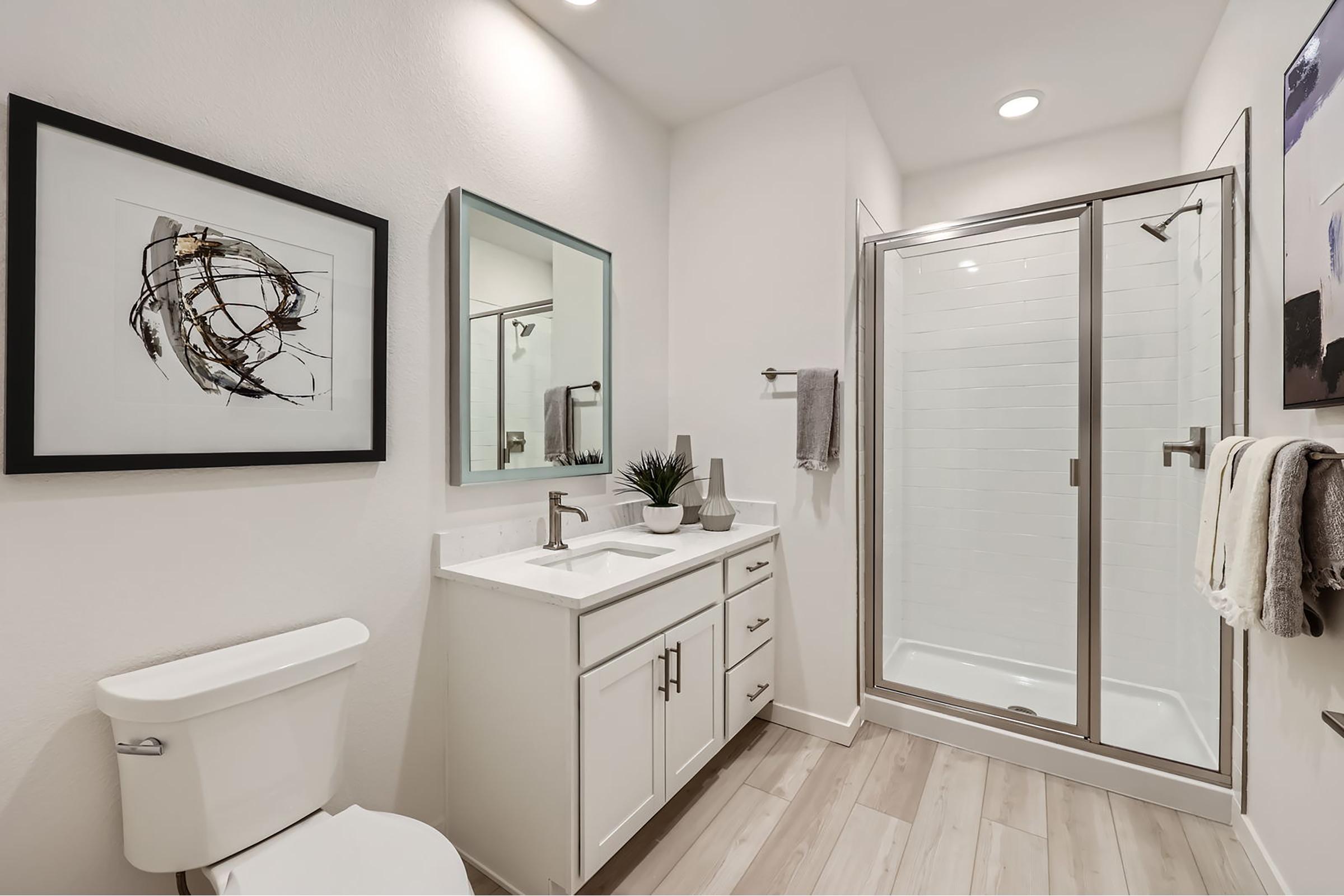 A modern bathroom featuring a white color scheme. The space includes a sleek toilet, a double-sink vanity with a mirror, and a glass shower. Decorative art hangs on the walls, and a small plant adds a touch of greenery. Light wooden flooring complements the contemporary design.