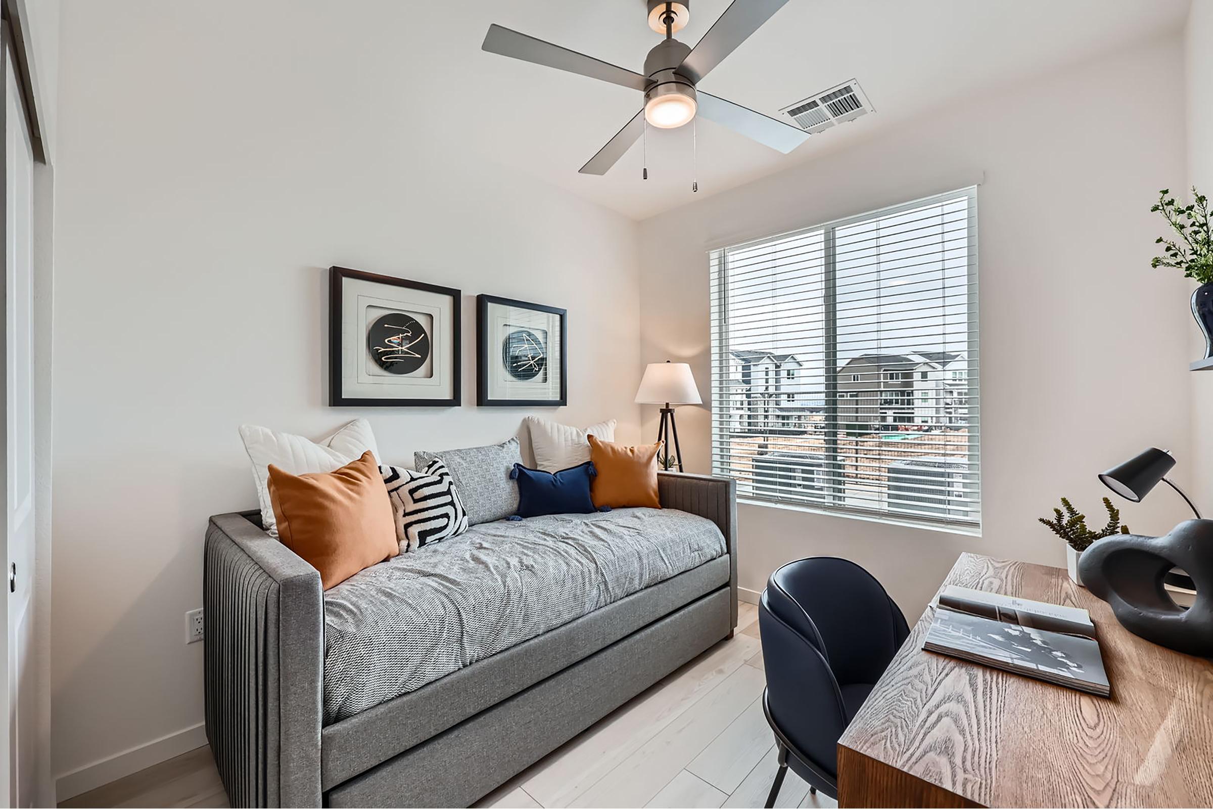 Cozy bedroom featuring a daybed with decorative pillows, a study desk with a chair, and a lamp. Natural light filters through a large window adorned with blinds, offering a view of an outdoor area. Modern wall art adds a touch of style to the minimalist decor.