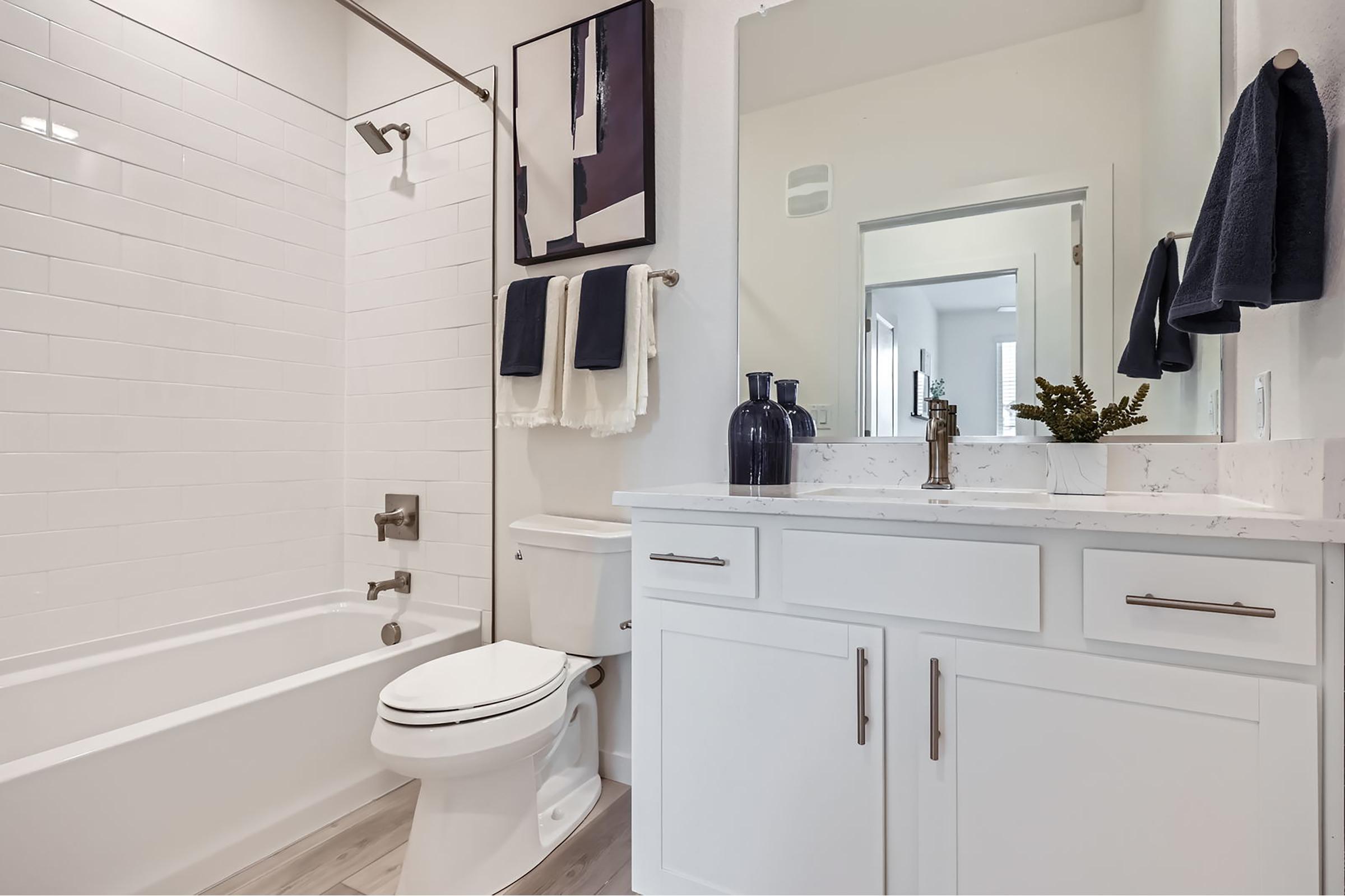 A modern bathroom showcasing a white tiled shower with a glass door, a white toilet, and a stylish vanity with a marble countertop. The vanity has a faucet and decorative items, while towels hang neatly on a rack. Natural light enters through an adjoining room, enhancing the space's clean, contemporary design.