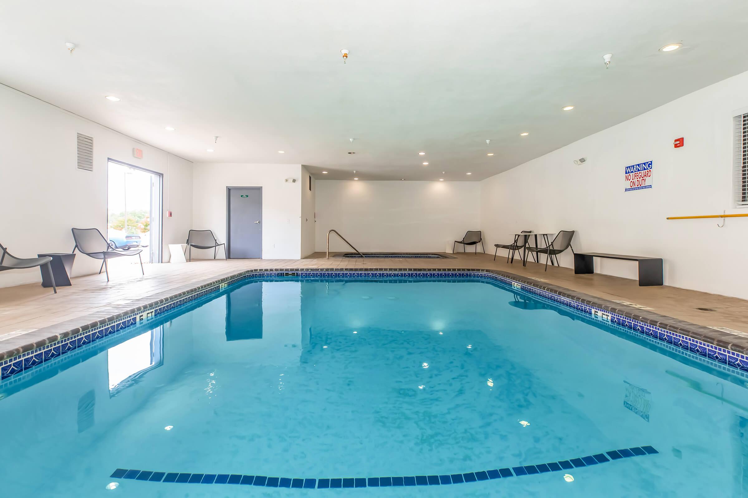 Indoor swimming pool area with clear blue water, surrounded by light-colored walls and a tiled floor. There are several black chairs arranged nearby and a small table. Bright overhead lights illuminate the space, creating a clean and inviting atmosphere.