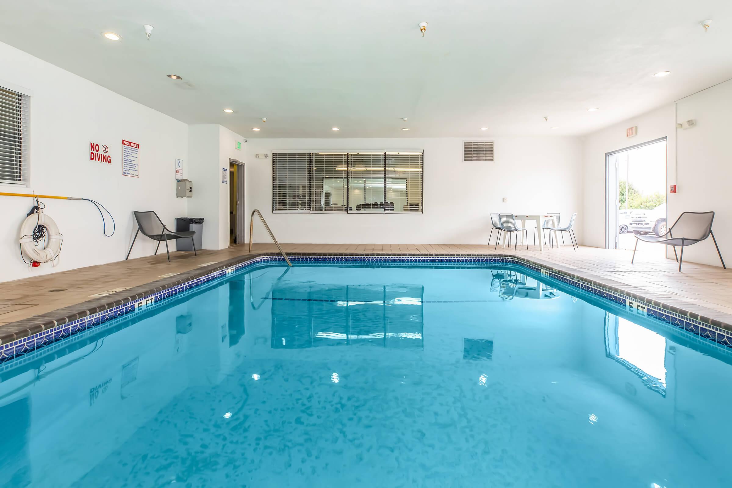 Indoor swimming pool with clear blue water, surrounded by beige walls. There are empty chairs placed around the pool area and a lifebuoy mounted on the wall. A large window allows natural light to enter, and there are signs indicating pool rules.