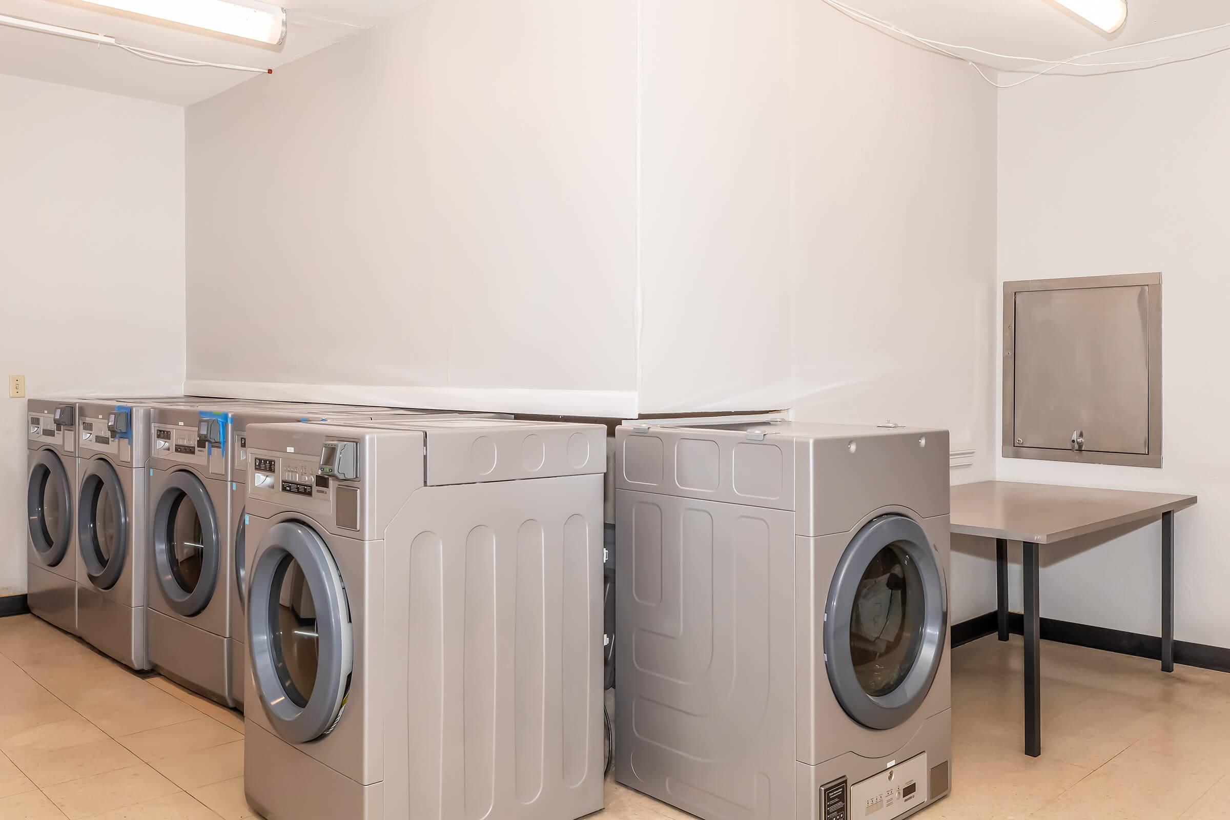 A clean, well-lit laundry room featuring four silver washing machines lined up against a wall. There is a gray table in the corner and a small utility sink mounted on the wall nearby. The floor is tiled and the walls are painted white.