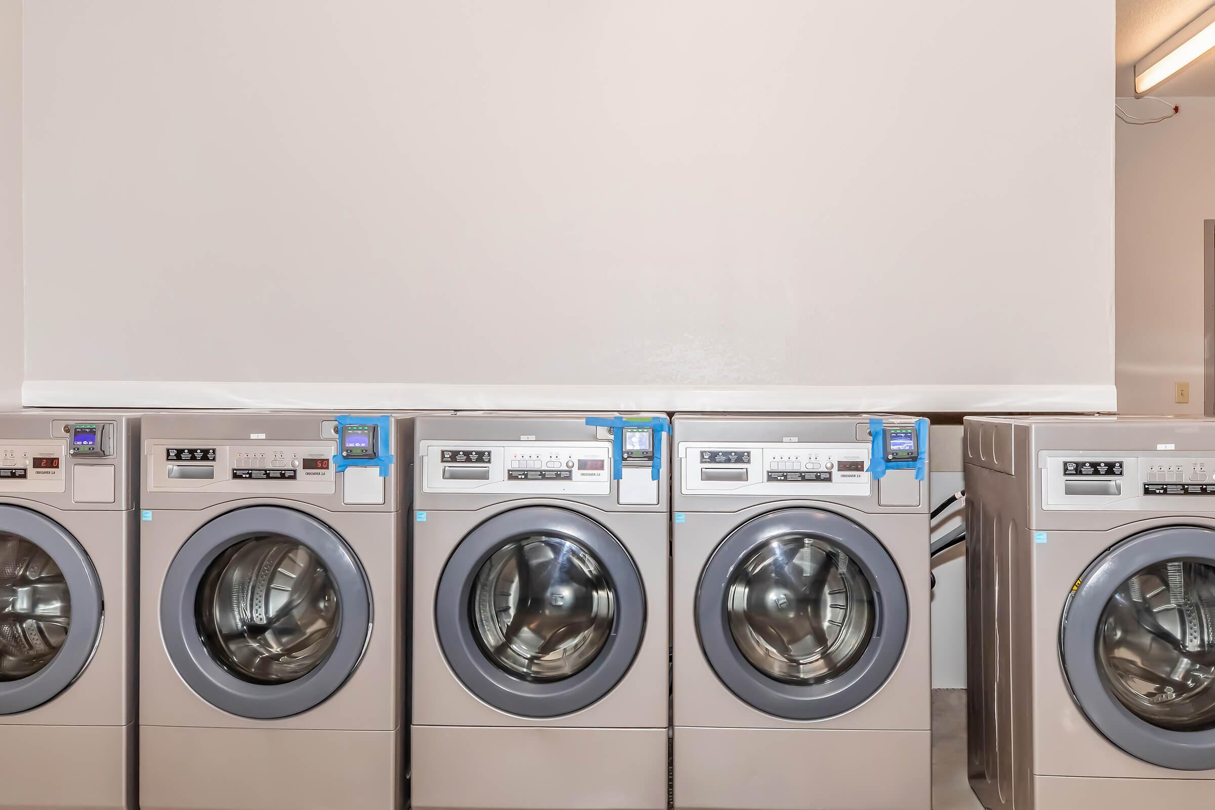 A row of modern, silver commercial washing machines with large glass doors, each equipped with control panels featuring buttons and slot for coins or cards. The machines are lined up against a plain wall in a well-lit laundry facility.