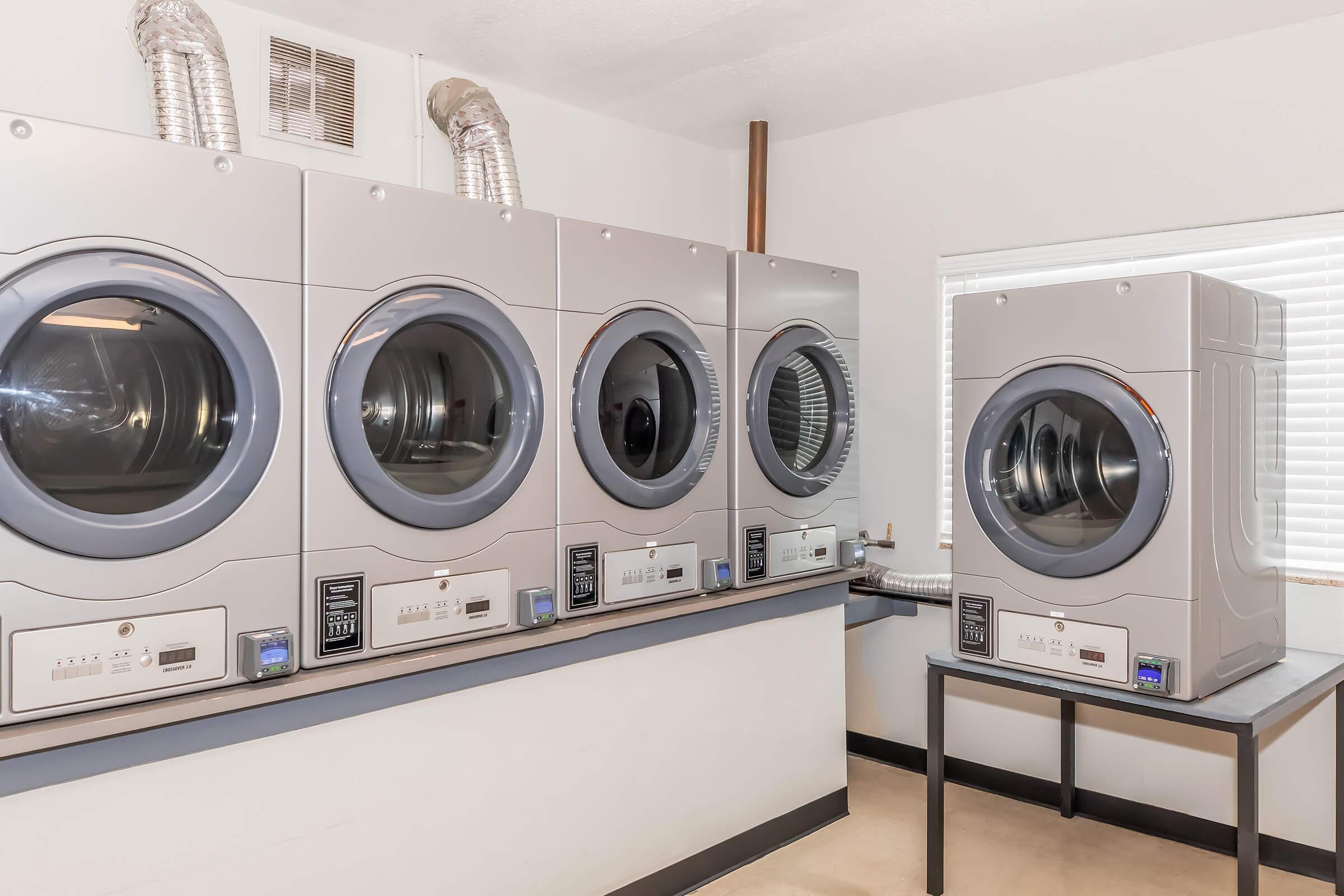 A row of modern washing machines lined up against a wall in a laundry room. Each machine features a large viewing window and control panel. Natural light streams in through a nearby window, illuminating the space. The room has a clean, organized appearance with a gray and white color scheme.
