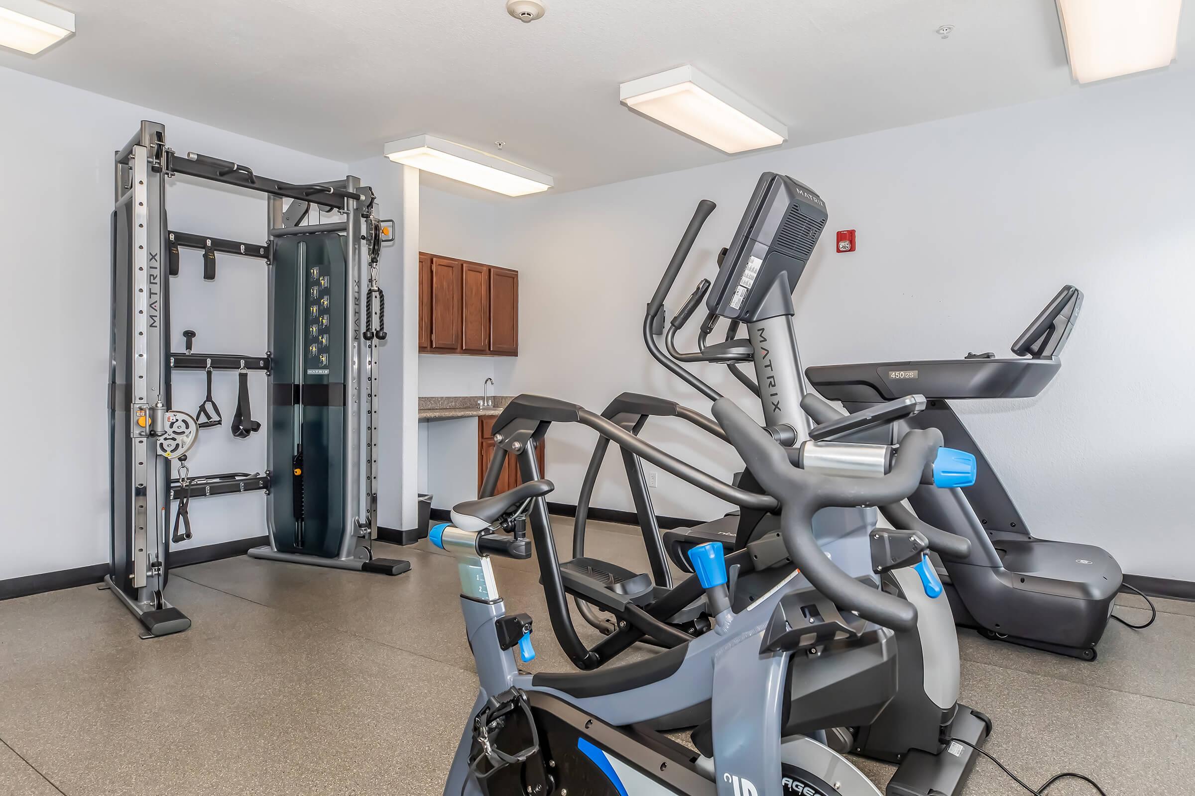 A well-equipped gym featuring various exercise machines, including a treadmill, an elliptical, and a multi-station weight machine. The spacious room has natural lighting, light-colored walls, and a countertop with cabinets in the background, creating an inviting workout environment.
