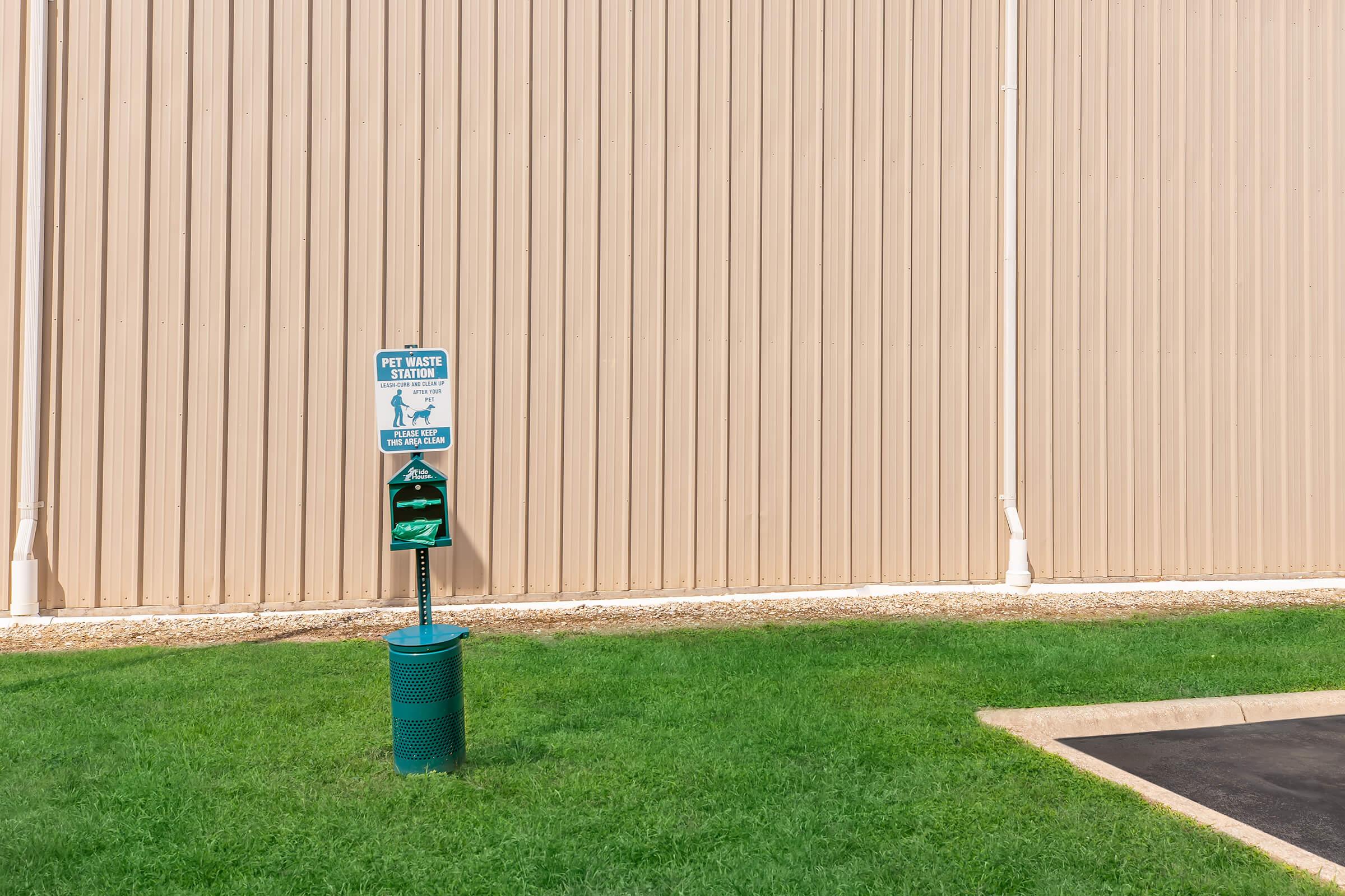 A green "Pet Water Station" sign mounted on a pole, next to a water dispenser for dogs. The station sits on a patch of grass in front of a beige corrugated metal wall.