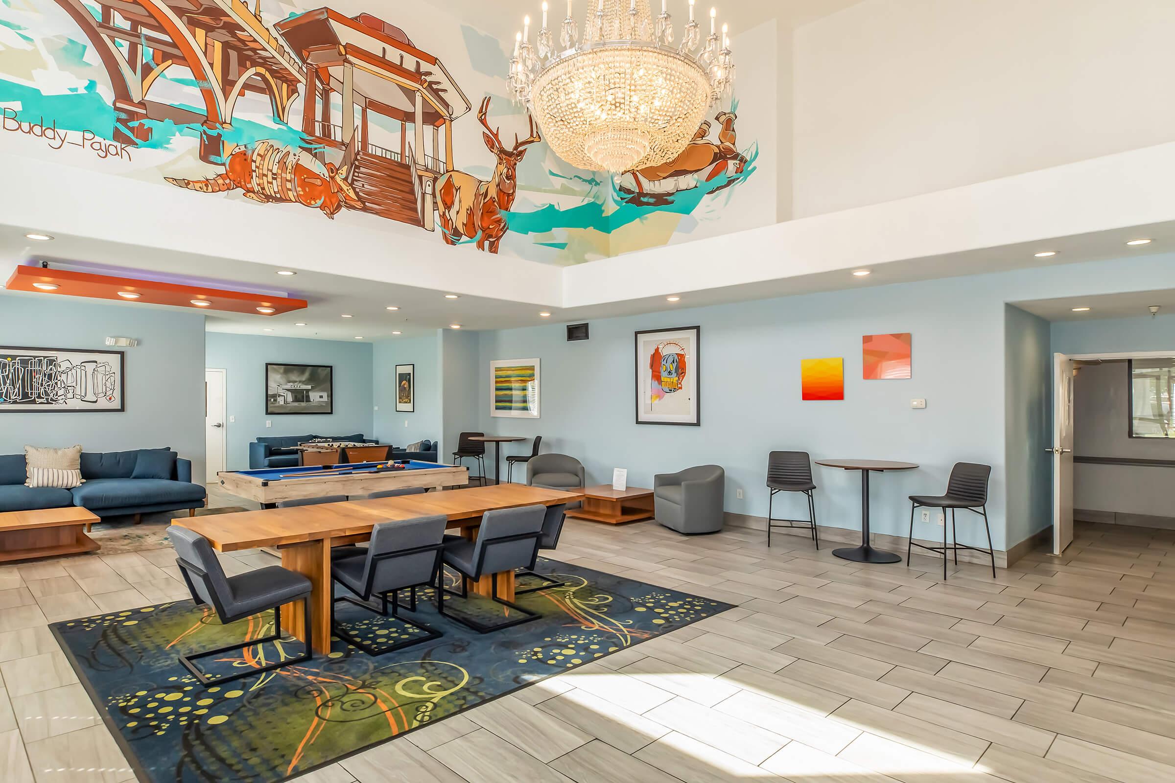A spacious and modern lobby area featuring a large wooden dining table surrounded by gray chairs, a comfortable blue seating area, and a colorful mural on the wall. A chandelier hangs from the ceiling, and large windows allow natural light to fill the space, highlighting the elegant decor and inviting atmosphere.