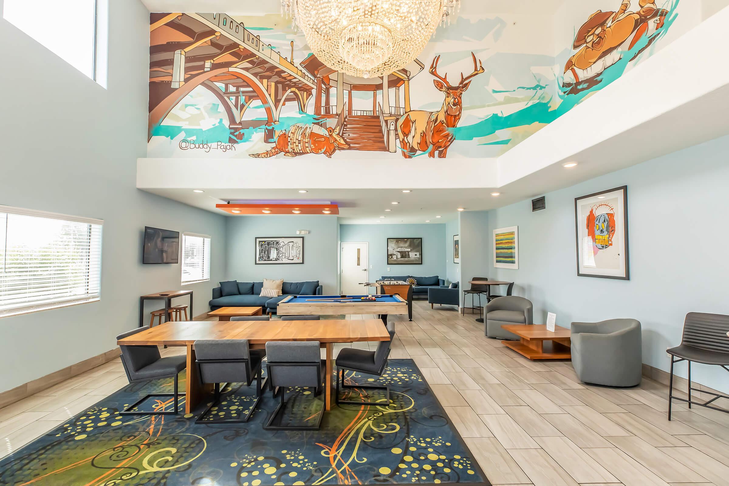 A modern interior of a spacious lounge area featuring a large chandelier, mural of animals on the wall, blue sofas, wooden tables, and stylish seating. Natural light fills the room through large windows, creating a welcoming atmosphere.