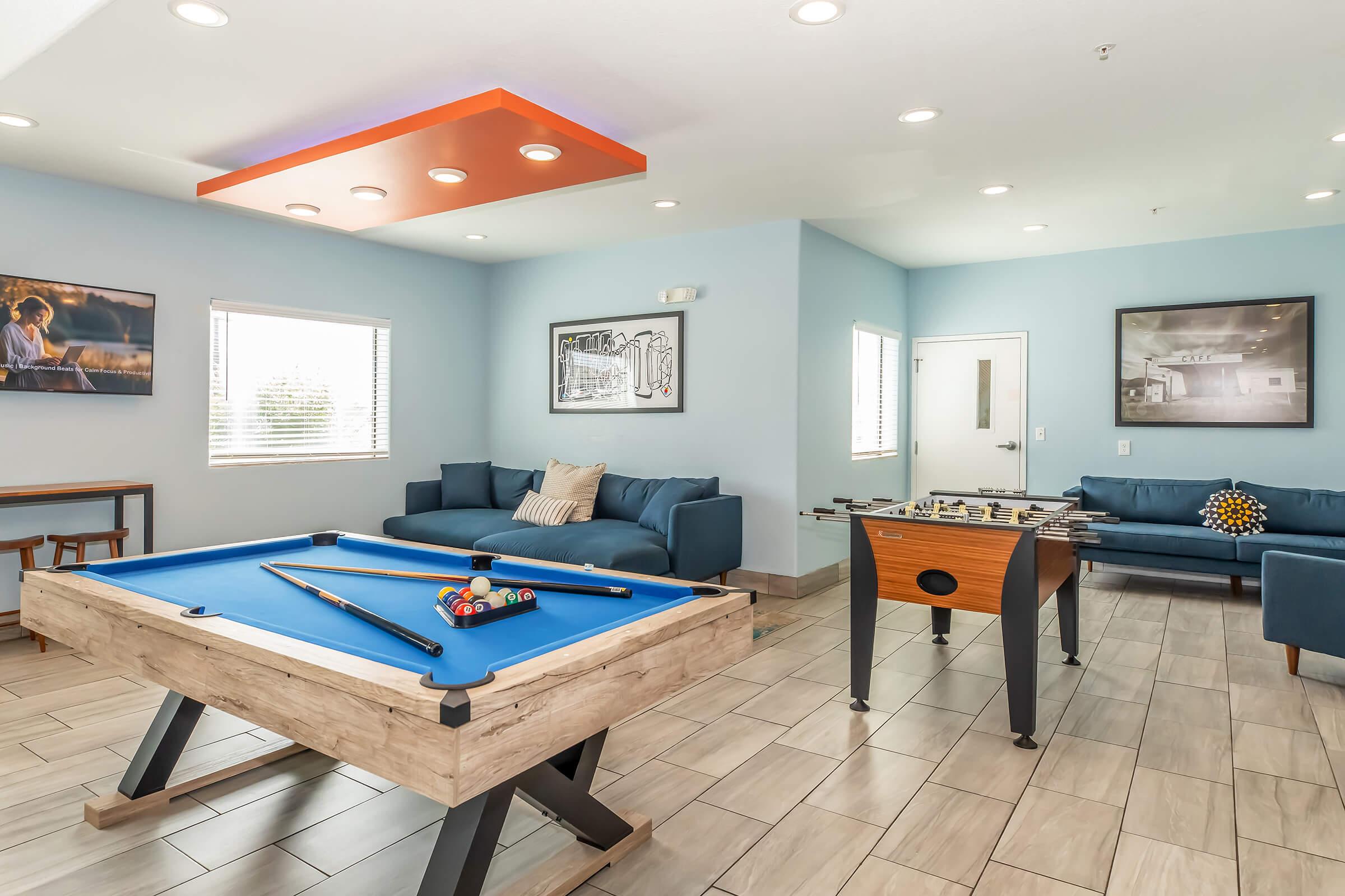 A bright game room featuring a blue pool table with cues and balls, a foosball table, and comfortable seating. The walls are adorned with framed artwork, and large windows allow natural light to fill the space. The ceiling has an orange accent section, enhancing the modern decor.