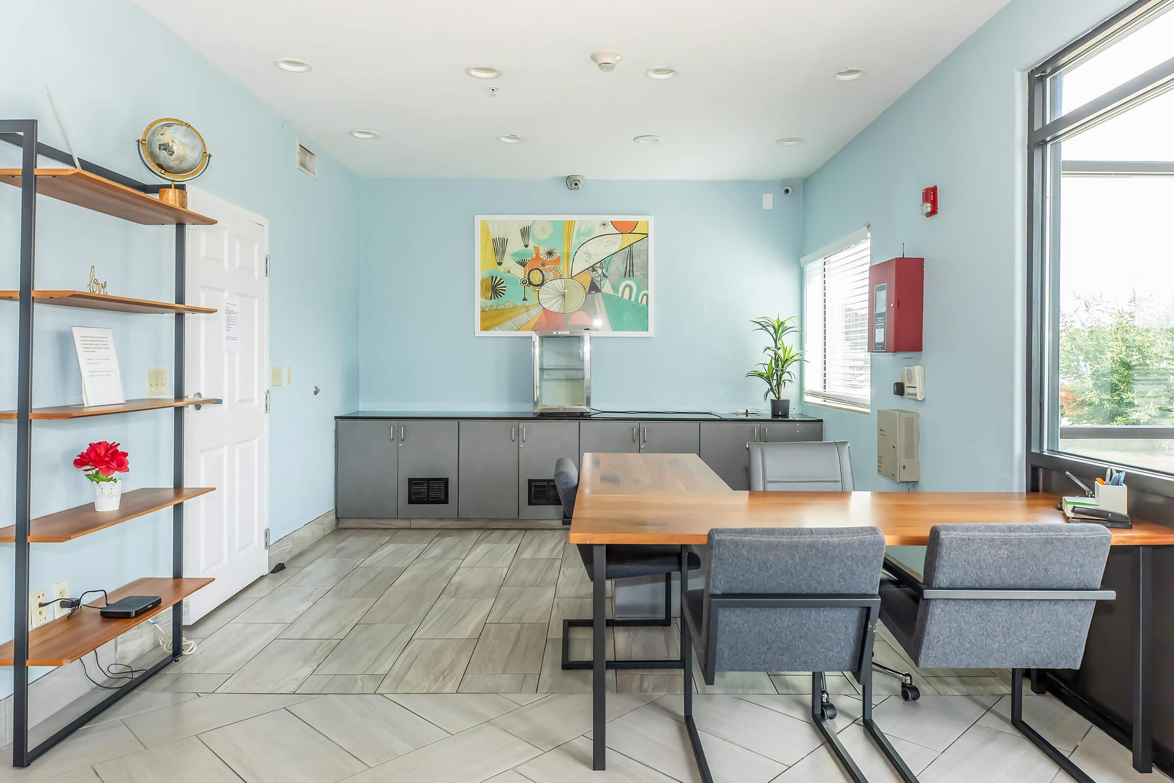 A modern office space featuring a light blue wall, a large window with outside views, a wooden table with gray chairs, and a stylish shelf with a decorative plant and art piece on the wall. The room has a clean, minimalist design with ample natural light.